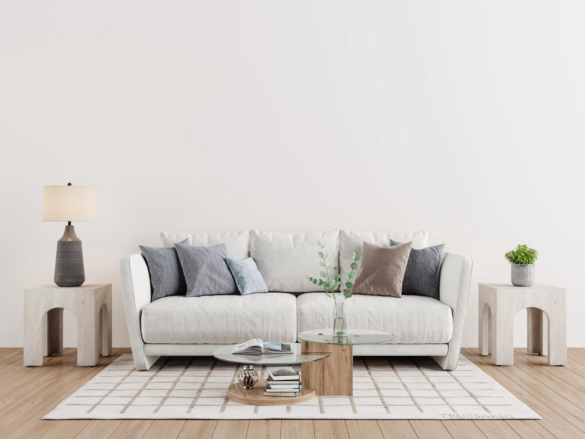Living room with a white sofa, blue and brown pillows, a beige and gray table lamp, and two white side tables with a potted plant and a glass coffee table holding books and decorative items, all on a light wood floor and beige rug.