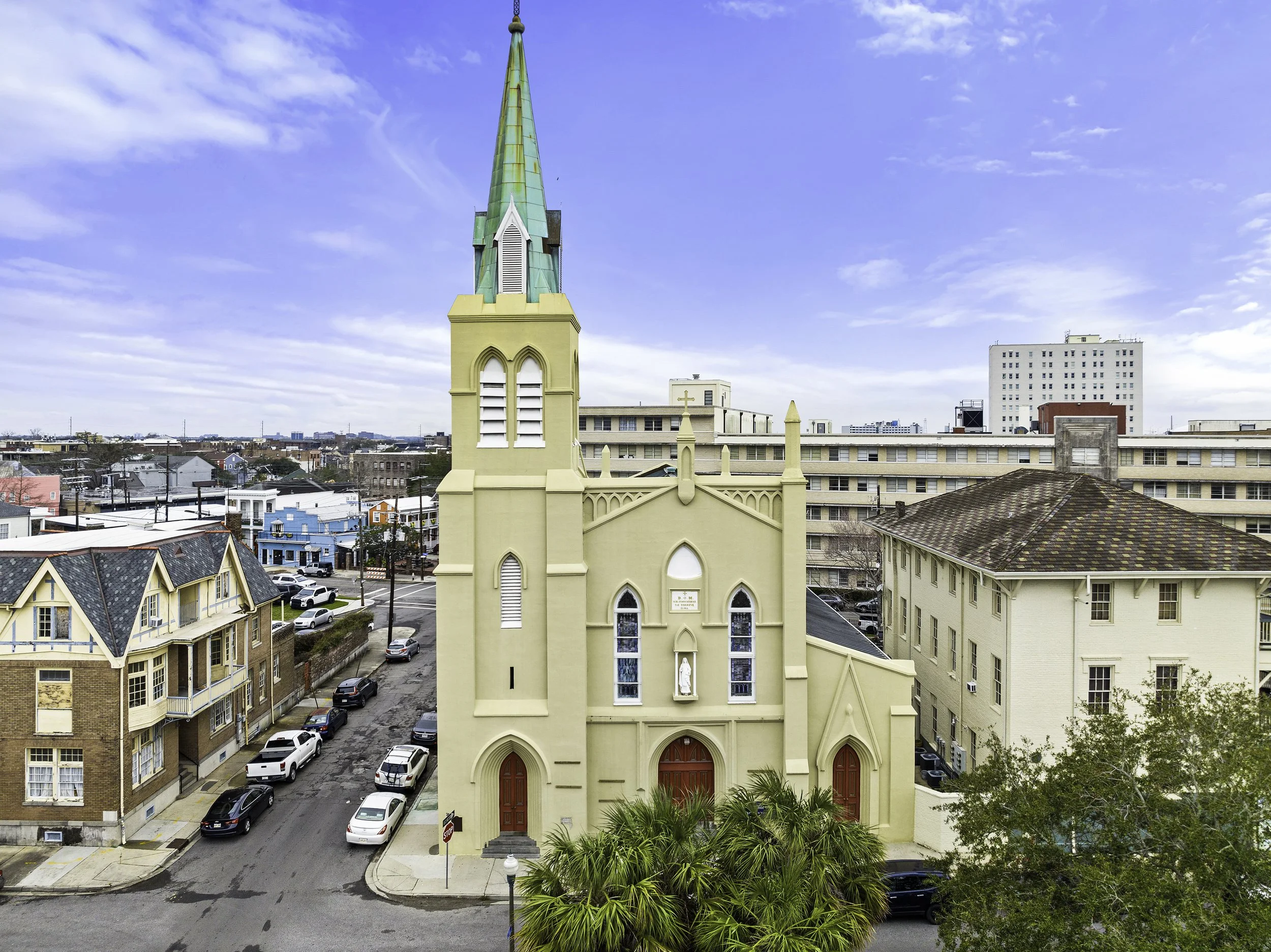 A yellow church with a green steeple and stained glass windows is in an urban setting with several other buildings and parked cars. The sky is partly cloudy.