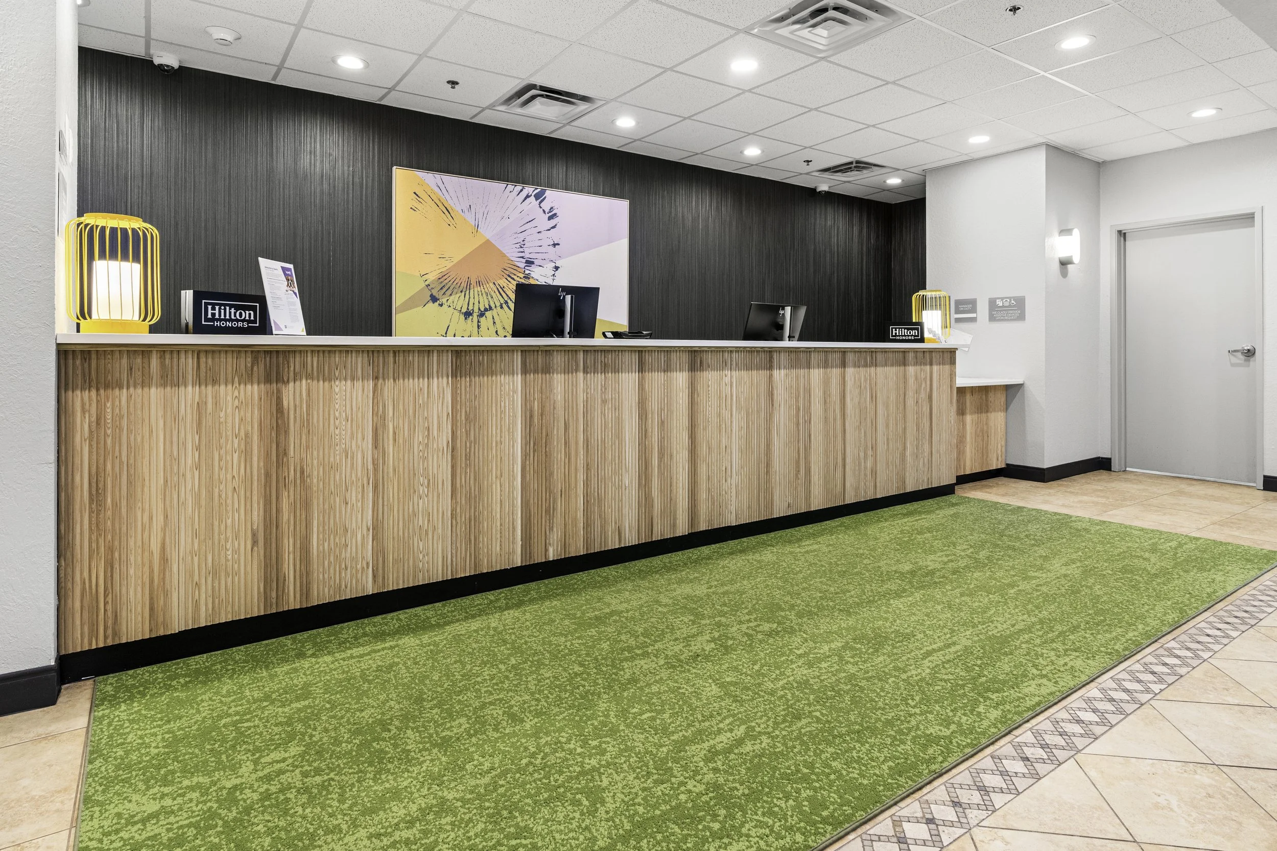 Hotel reception area with a curved wooden counter, black computers, a colorful abstract artwork on a dark wall, yellow desk lamps, and green carpet flooring.