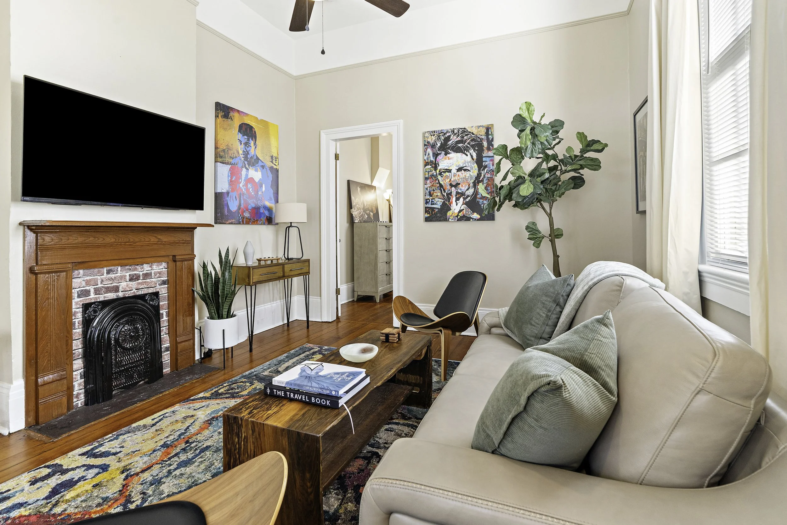 Living room with a beige sofa, a wooden coffee table with books, and a patterned area rug. There is a fireplace with a wooden mantel and a colorful portrait on the wall. A large potted plant is next to the sofa. The room has a ceiling fan and windows