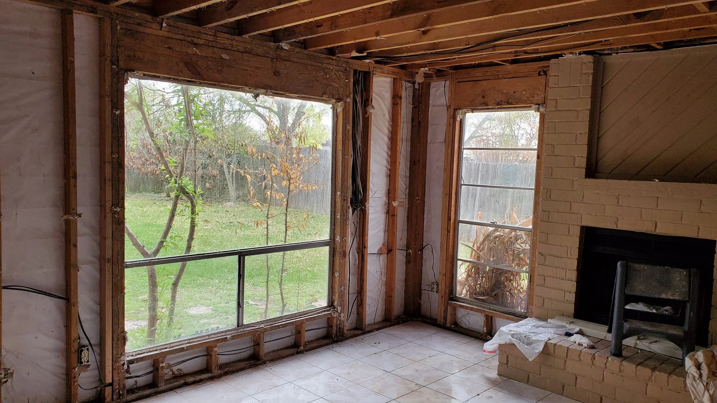 Living room under renovation with exposed wooden framing, two large windows overlooking a yard, and a brick fireplace with a black fire box. Tiled floor with some construction materials and a chair.