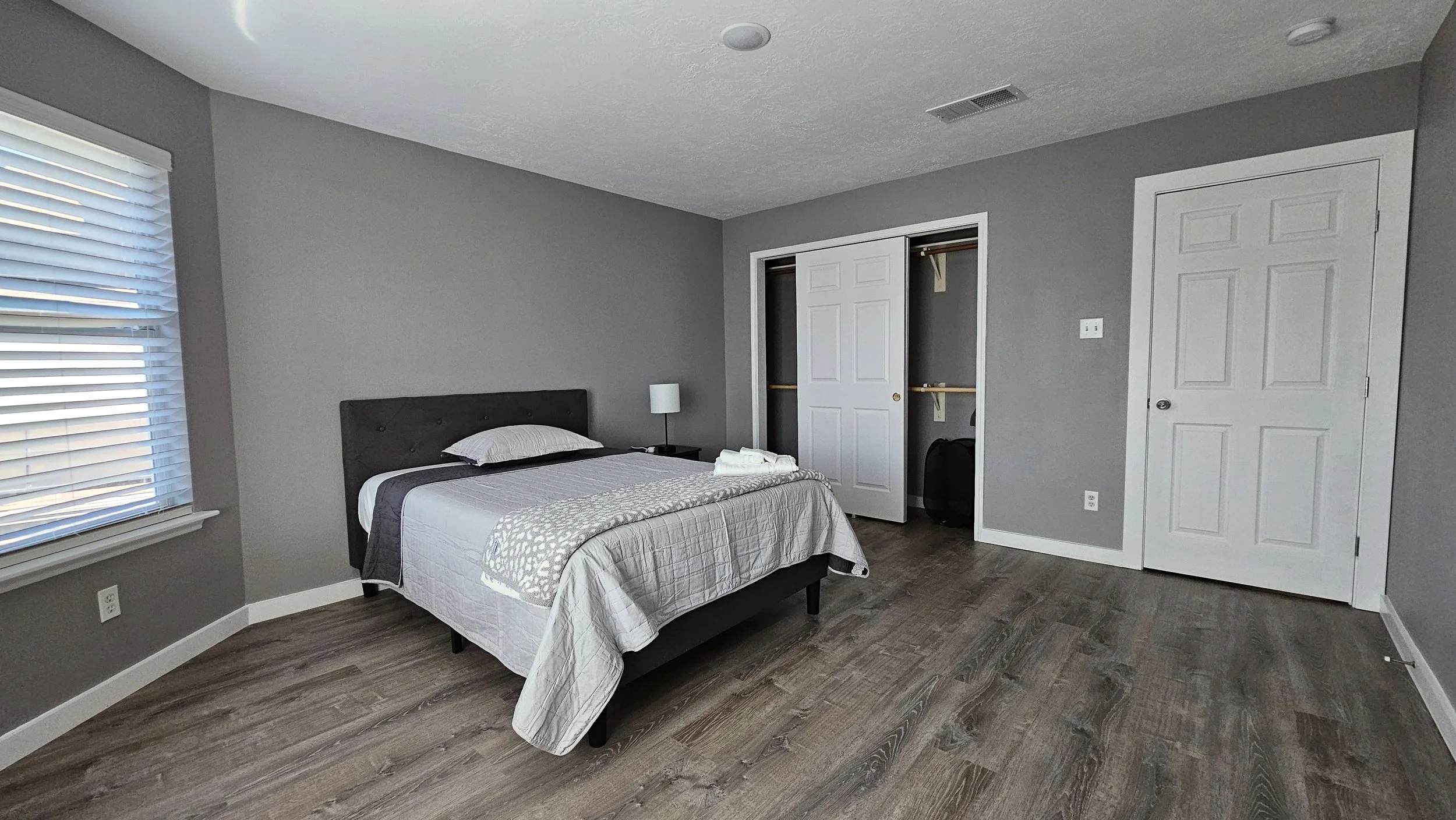 A bedroom with gray walls, a bed with white and gray bedding, a small bedside table with a lamp, an open closet with sliding doors, and a window with white blinds.