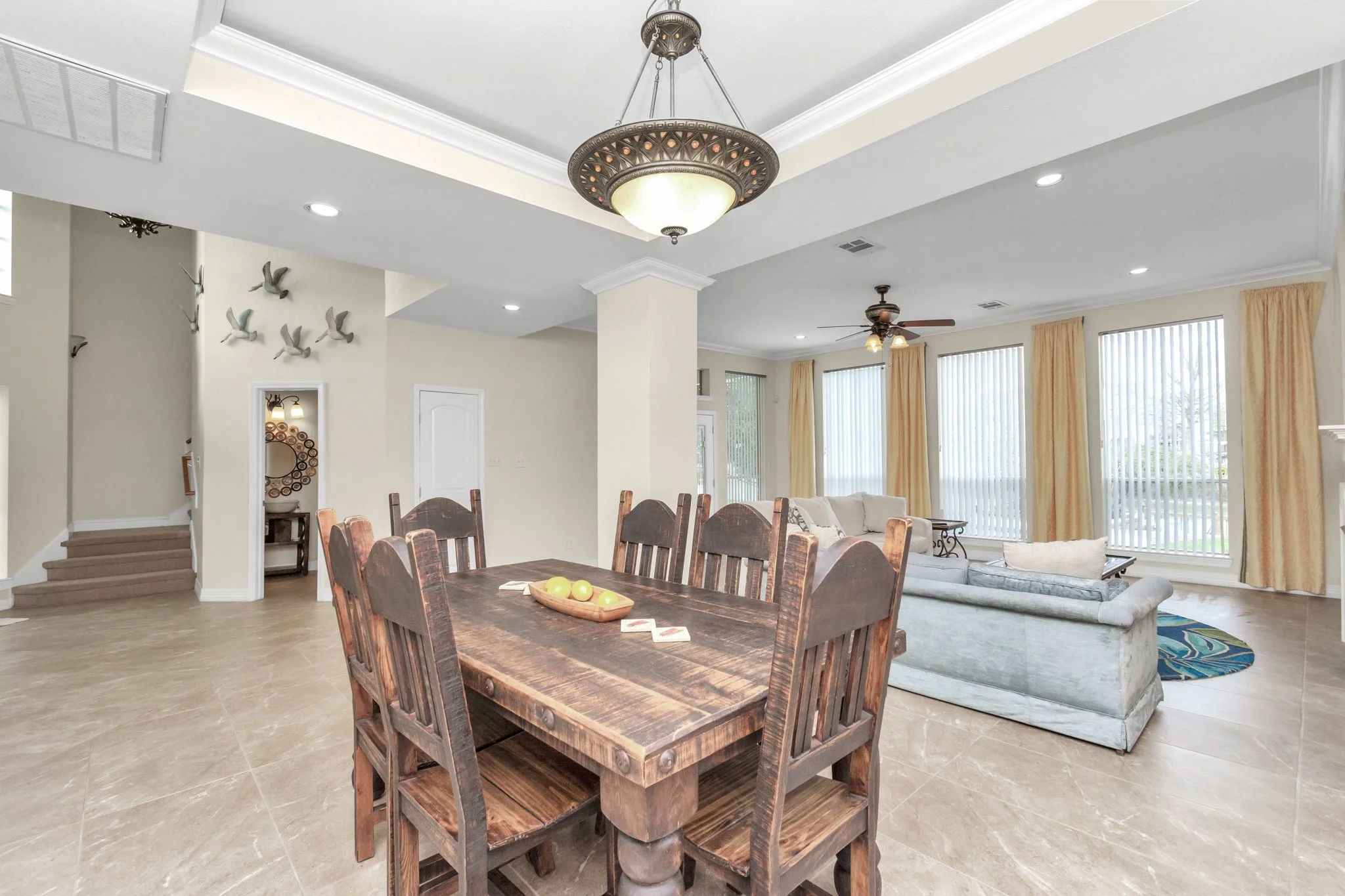 Open-concept living and dining room with wooden dining table, beige sofa, large windows with vertical blinds and yellow curtains, ceiling fans, chandelier, and wall decorations of flying birds.