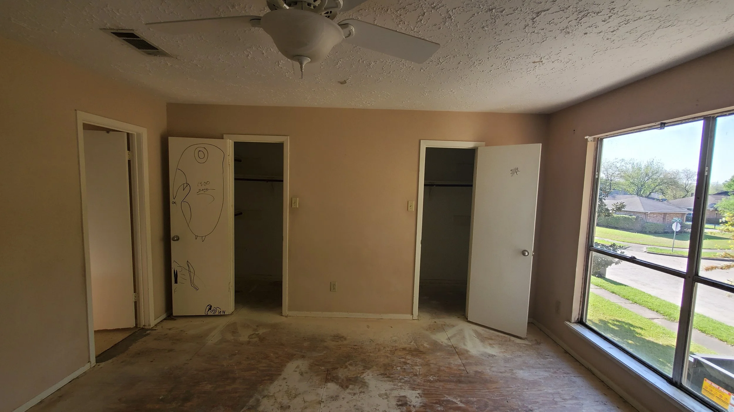 Empty room with a large window showing a view of a neighborhood with houses and trees. The room has beige walls, two open closets, a ceiling fan, and a worn floor.