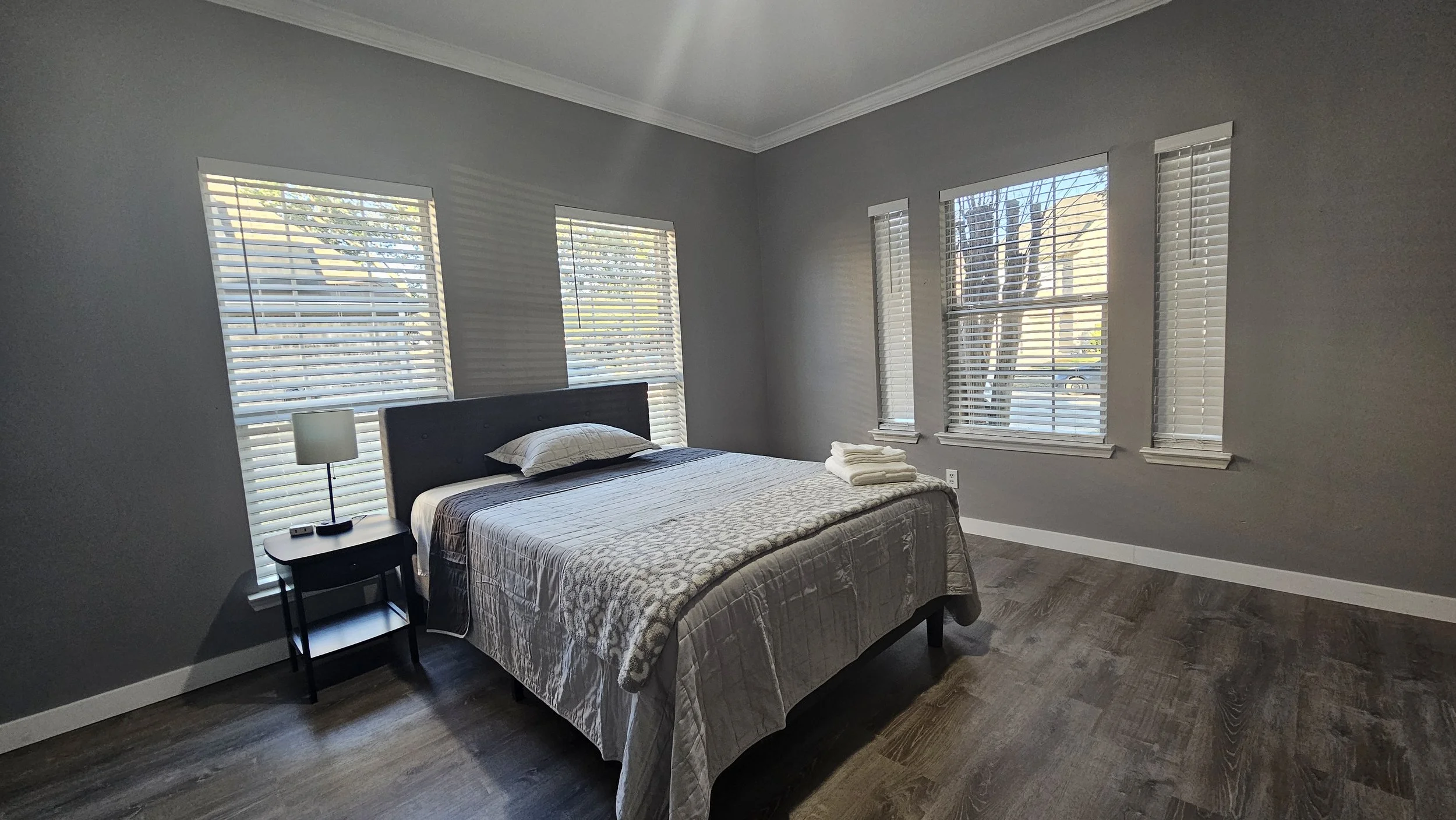 A minimalist bedroom with gray walls, hardwood floors, a black bed frame, a white and beige quilt, a pillow, a towel on the bed, a black nightstand with a lamp and TV remote, and five windows with white blinds allowing sunlight to enter.