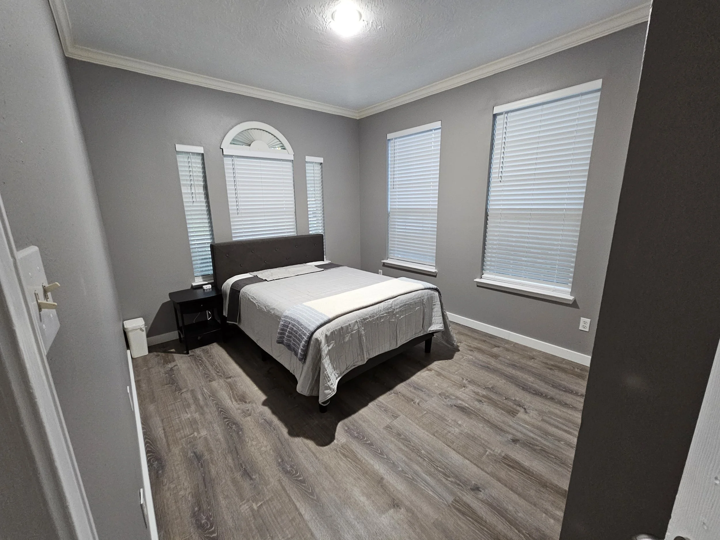 A minimalist bedroom with three windows covered by white blinds, a gray upholstered headboard on the bed, a white bedspread with a gray blanket at the foot, a small black side table with a phone and remote, light wood flooring, and crown molding on t