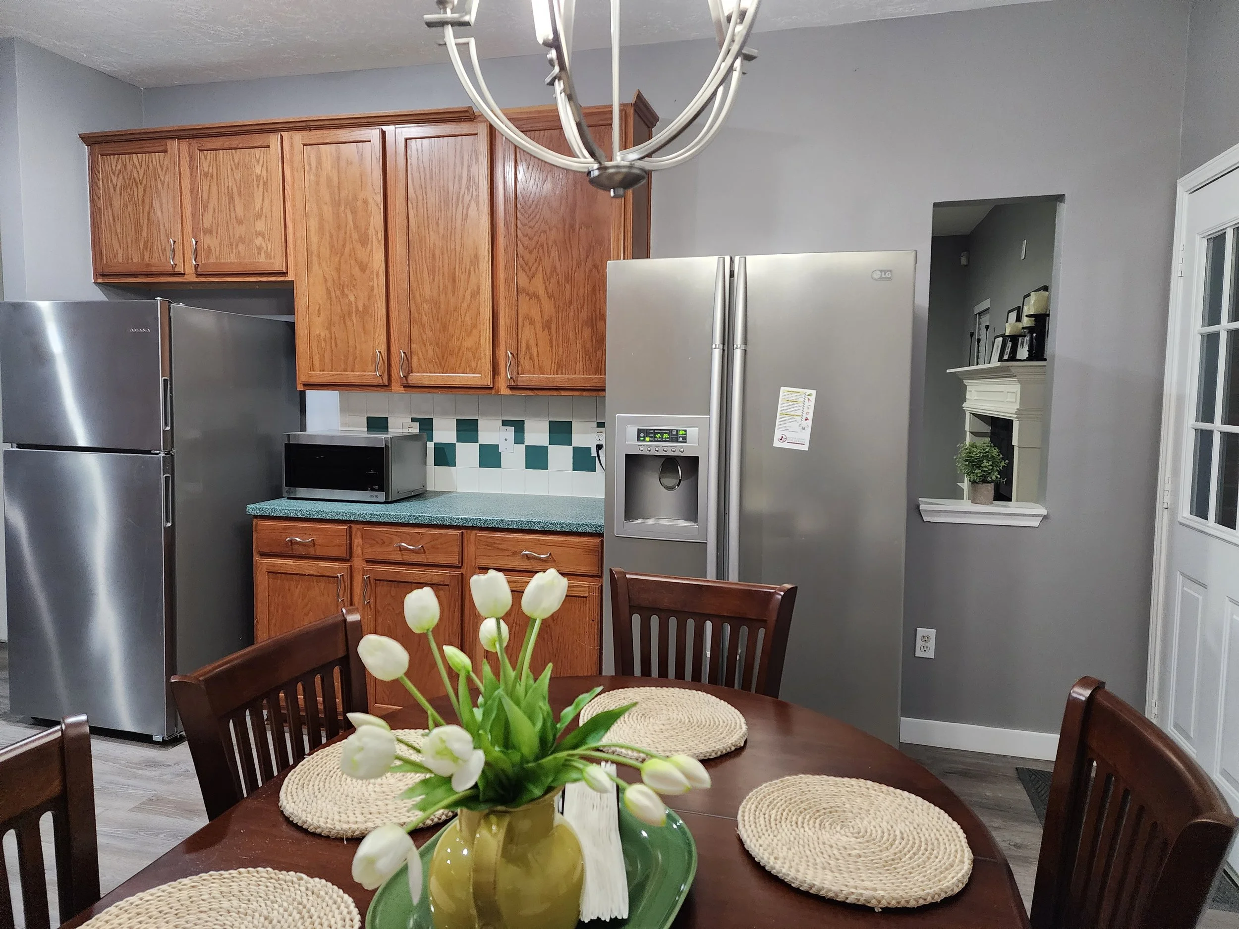Kitchen with wooden cabinets, stainless steel refrigerator, microwave on countertop, dining table with woven placemats, vase with white tulips, and a wall with small shelf and framed pictures.