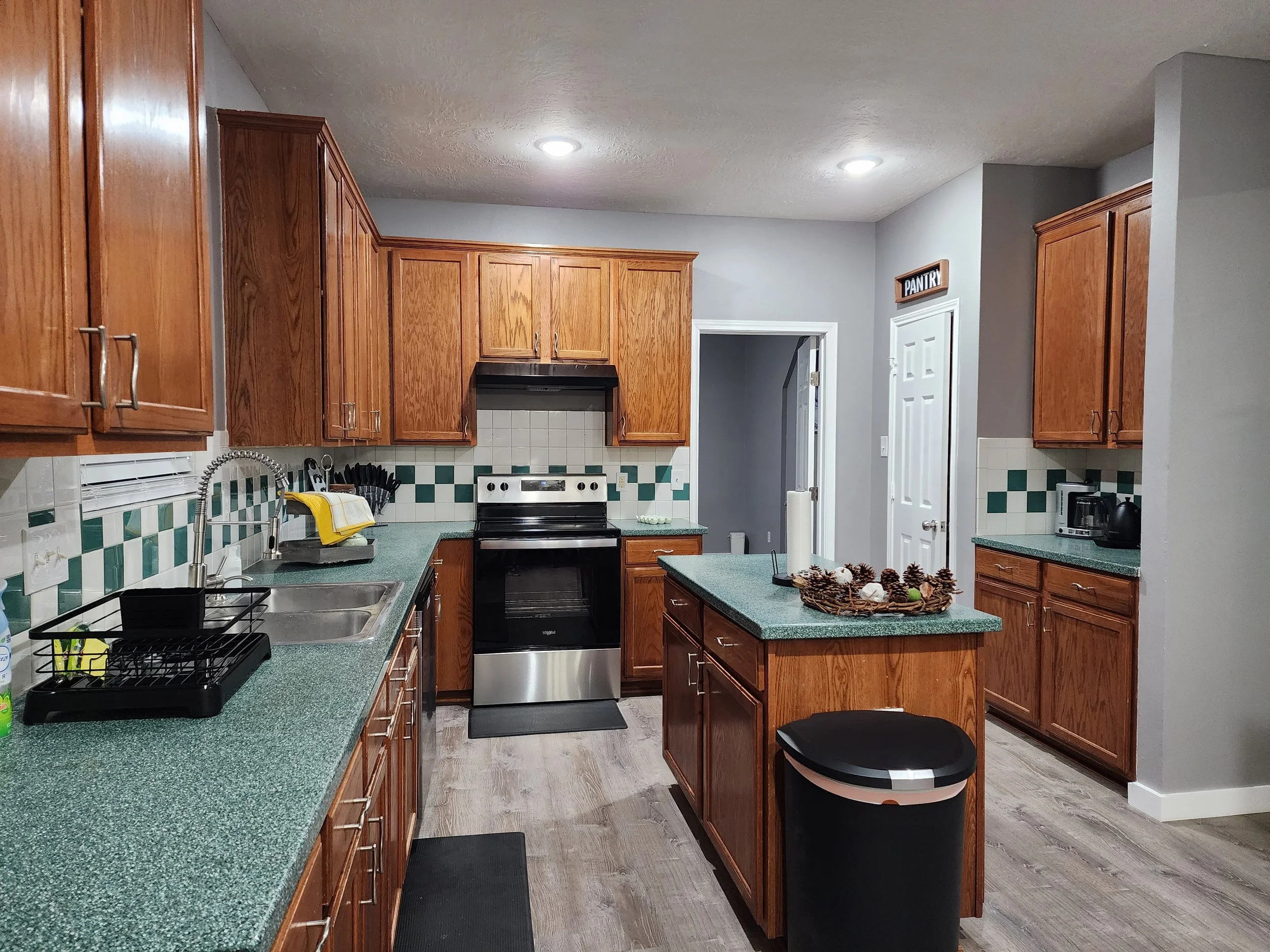 Kitchen with wooden cabinets, green countertops, a stove, sink, trash can, and small island in the center with a decorative centerpiece.