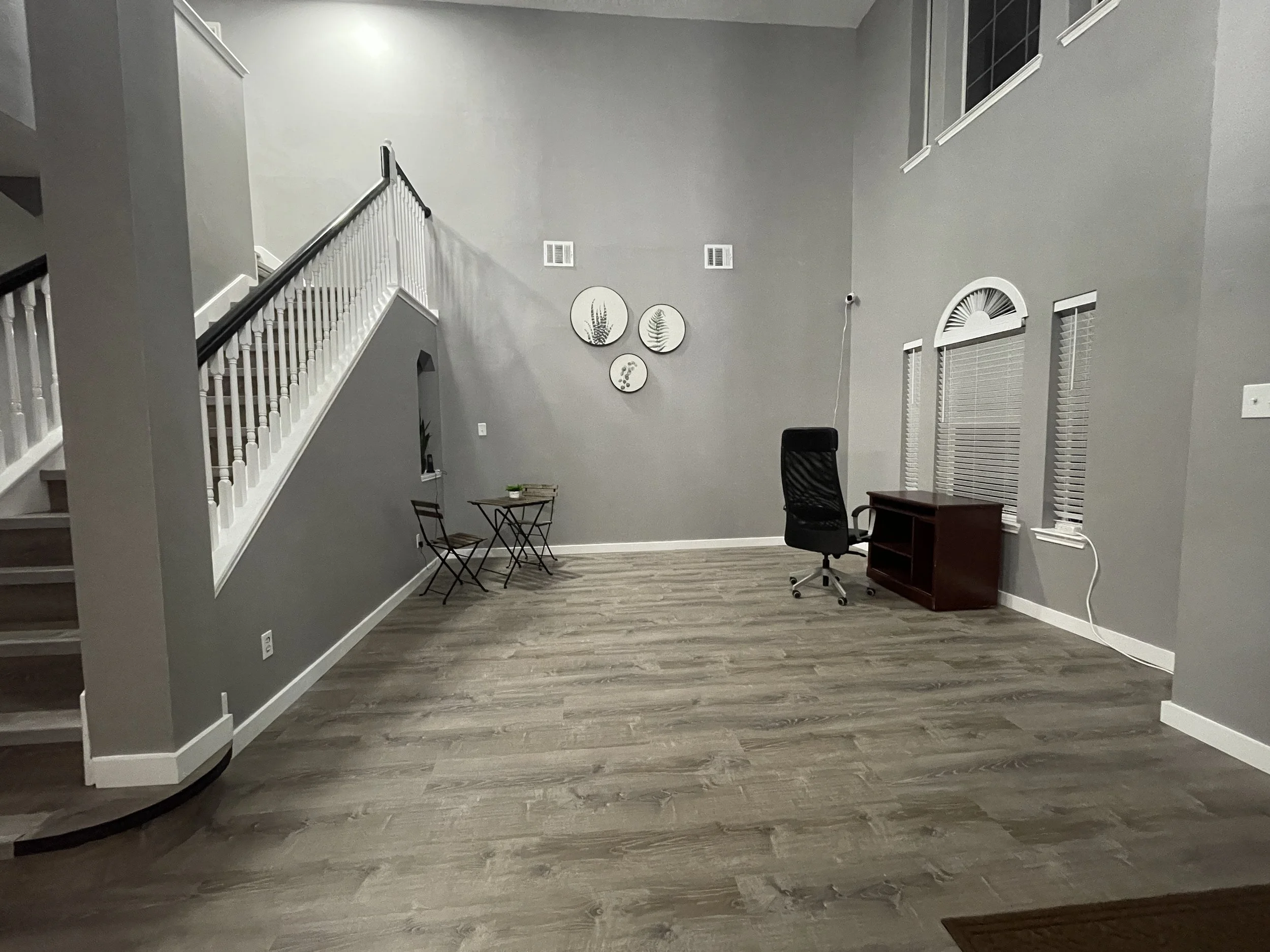 Empty living room with grey walls, wood flooring, a staircase on the left, a small round table with two chairs on the left side, a black office chair, a small wooden desk, three decorative wall plates with plant designs on the back wall, and several 