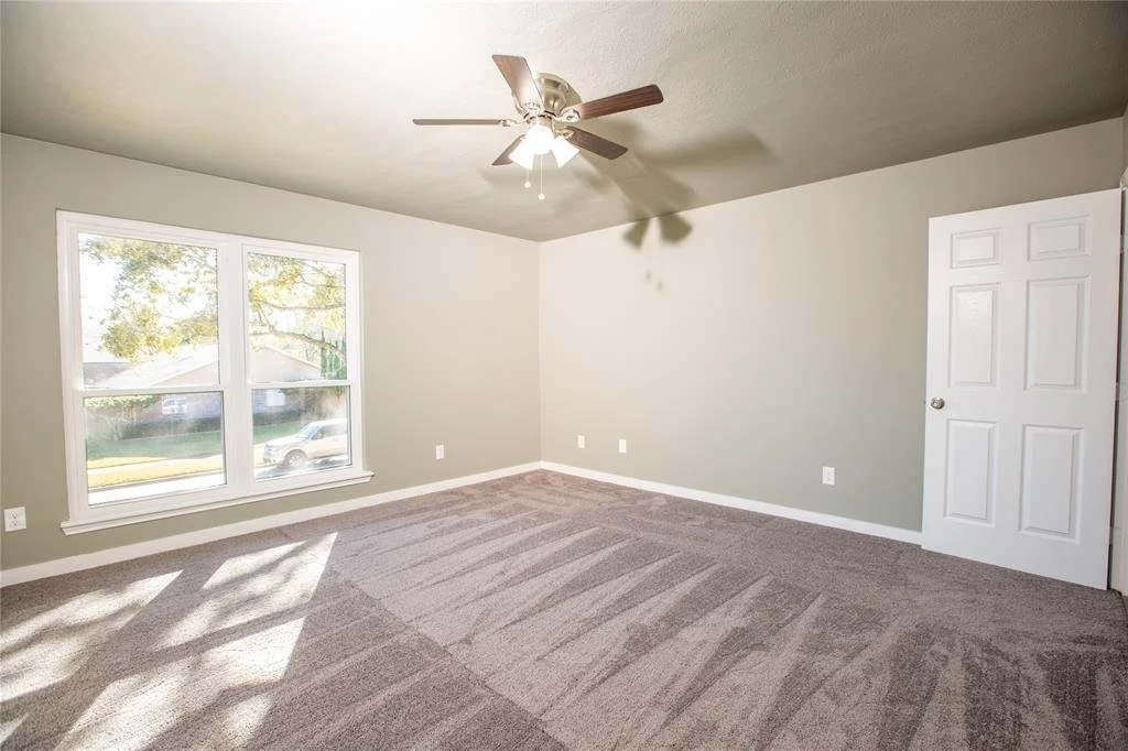 Empty living room with beige carpet, light green walls, a large window, and a ceiling fan with light.