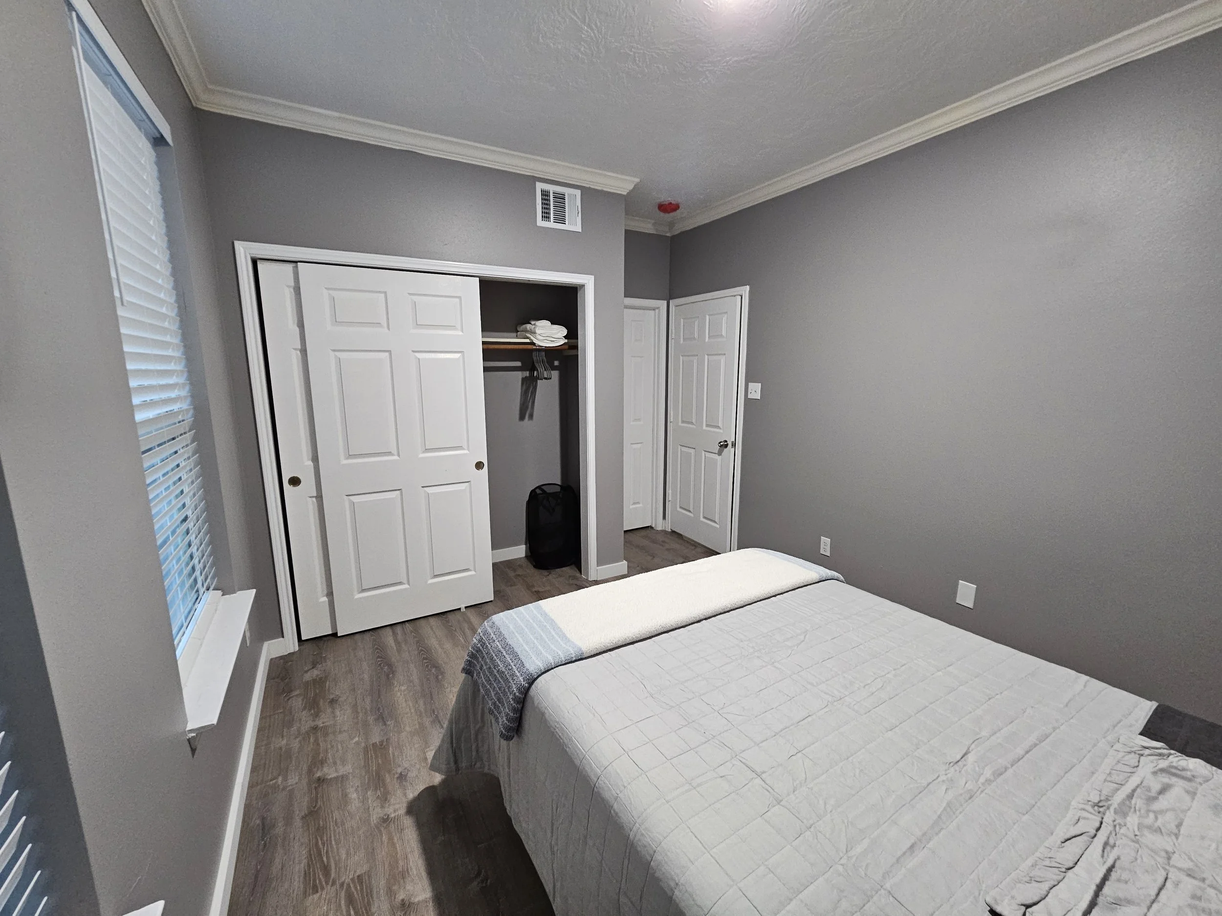 A bedroom with grey walls, a window with blinds, a bed with a grey and white quilt, a closet with white double sliding doors, a black suitcase inside the closet, and a wooden floor.