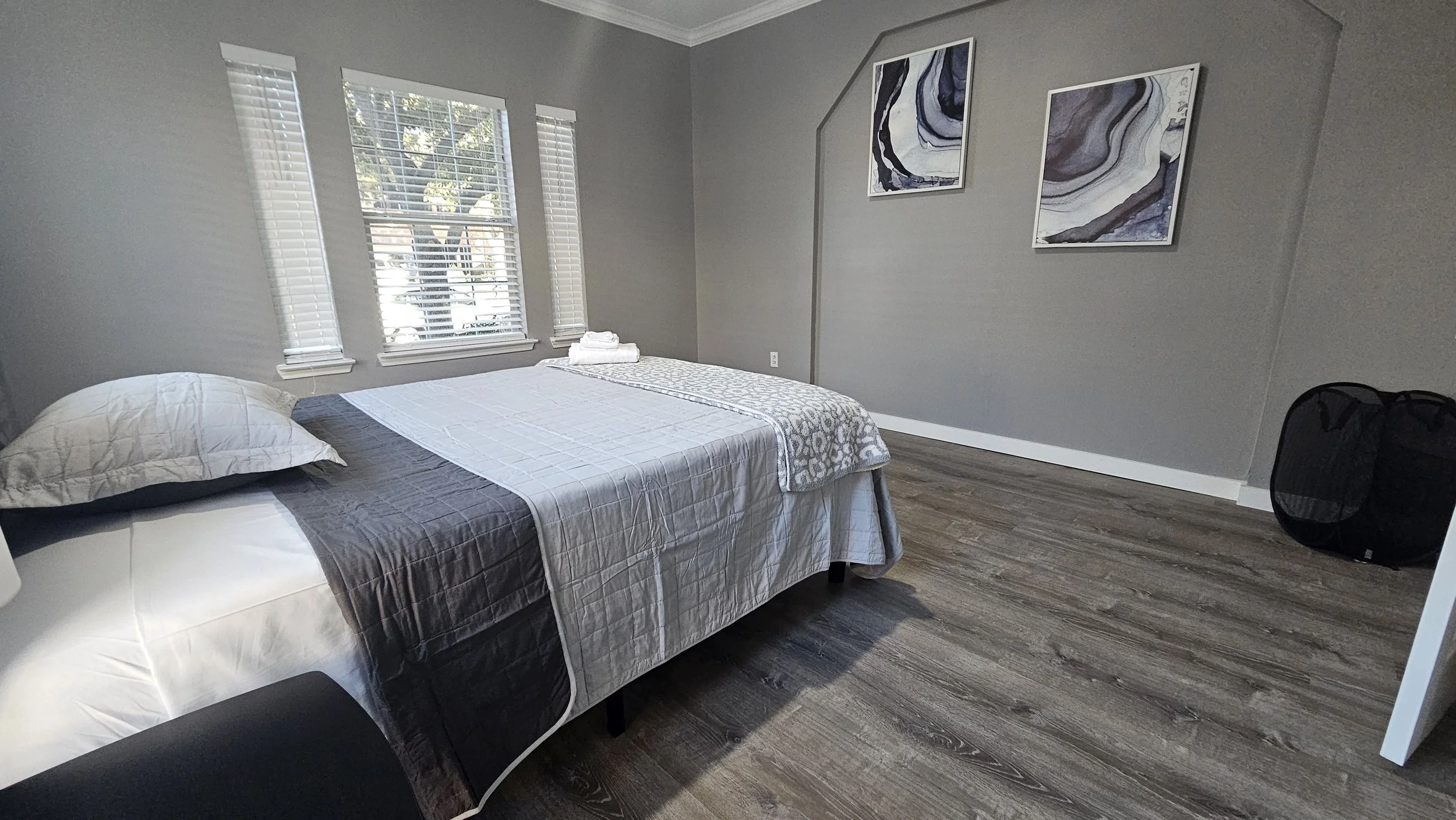 A bedroom with a large bed covered with a quilted gray and white bedspread, a pillow, and a gray blanket, positioned against a wall with three windows in the background, framed abstract artwork on the wall, and a black laundry hamper in the corner.