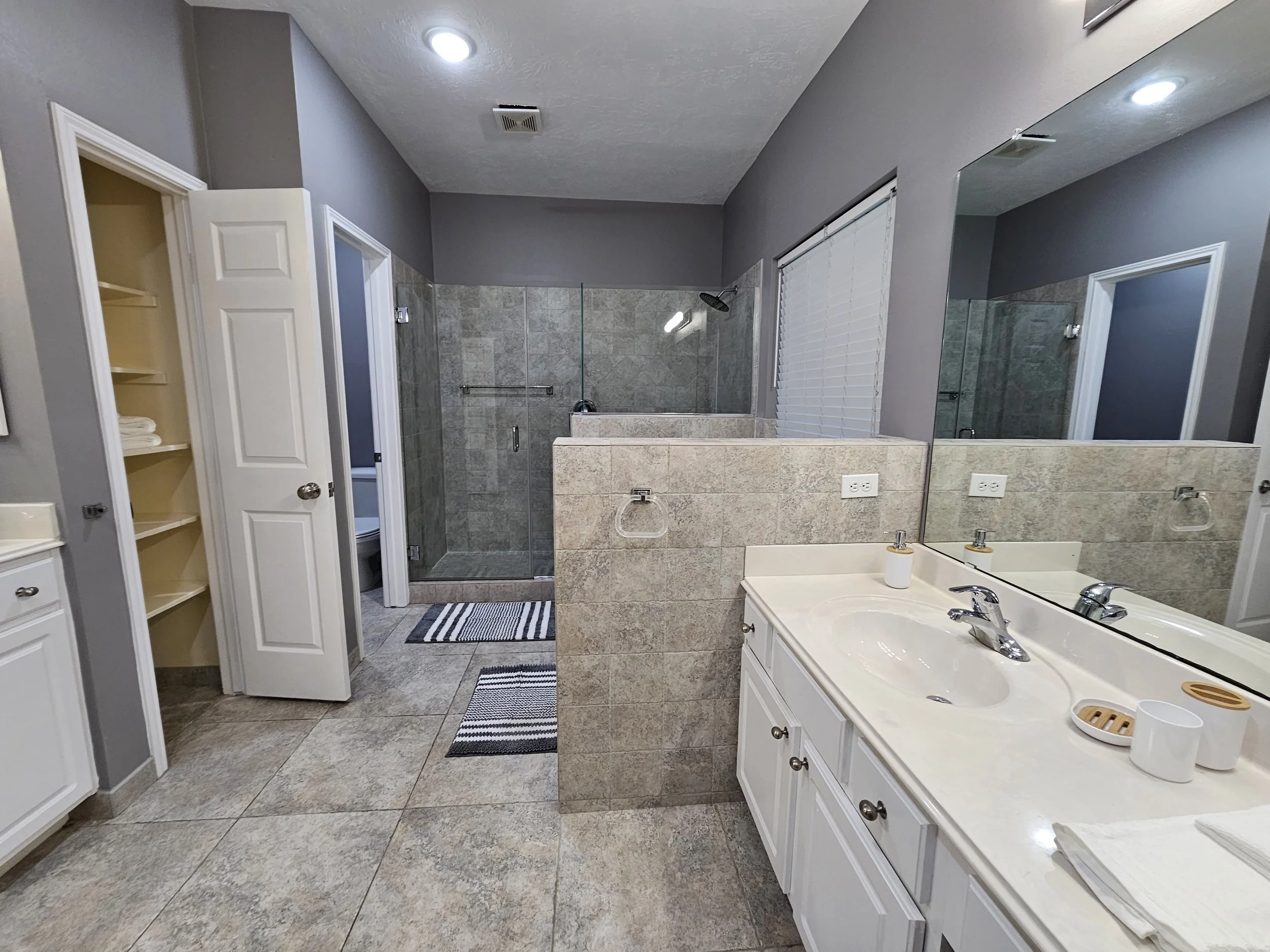 Bathroom with a vanity, sink, mirror, walk-in shower, and open storage shelves. Gray walls, large tiled floor, and a small window with blinds.