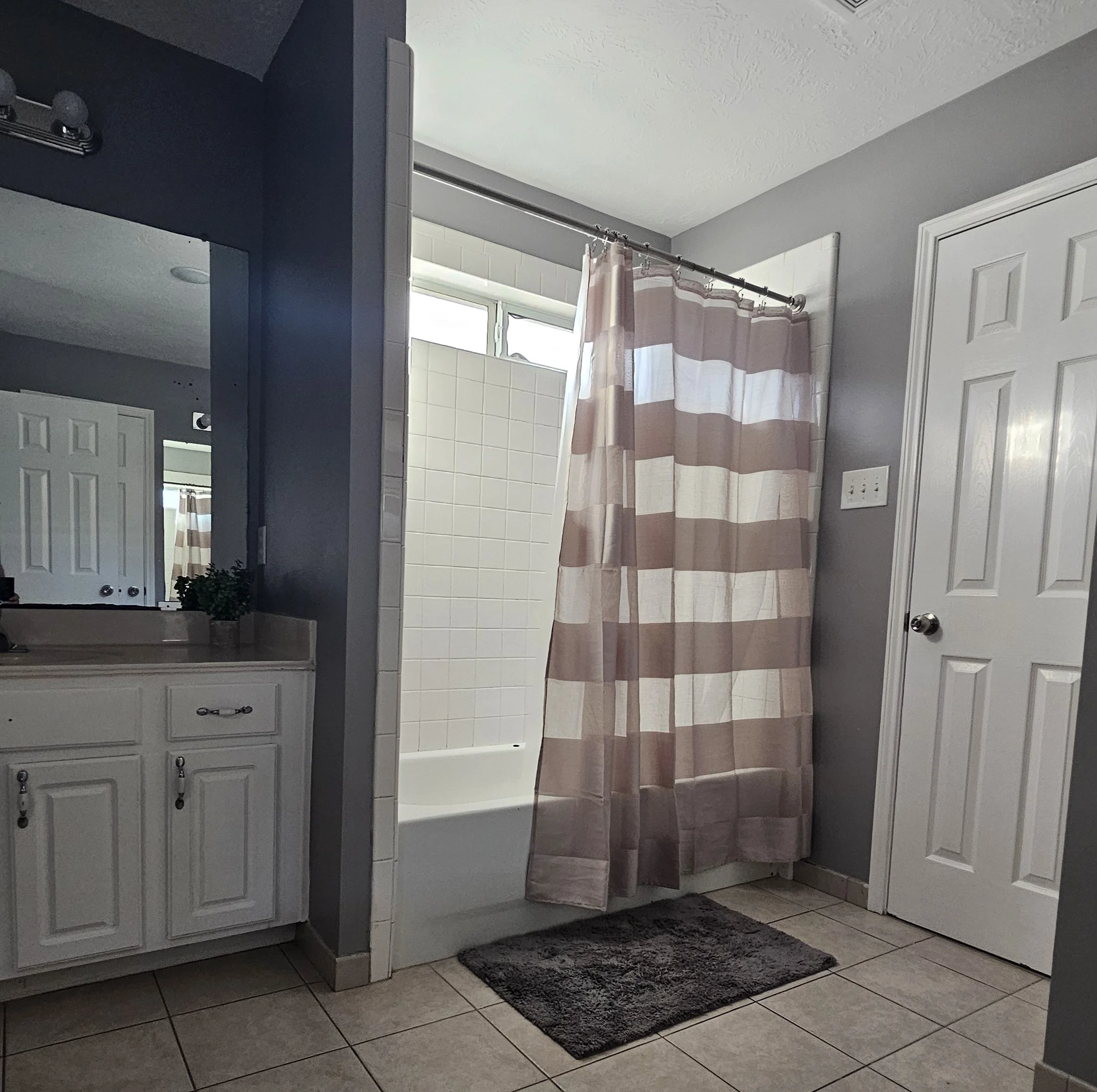 A bathroom with a shower behind a striped pink and white curtain, a white vanity with a mirror and small potted plant, tiled floor, and a white door.