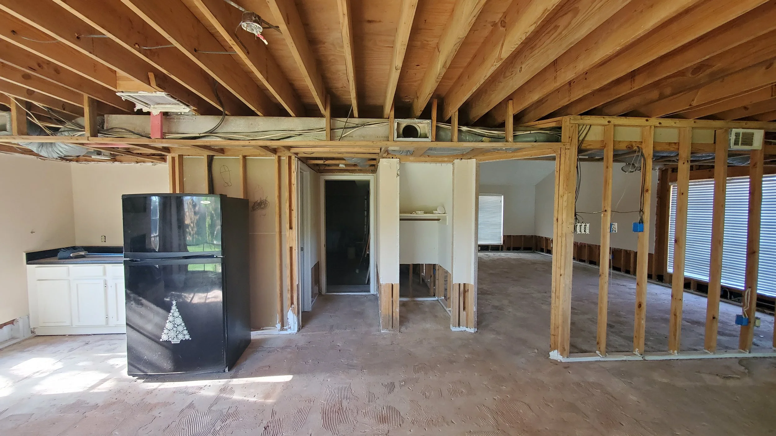 Interior of a house under renovation with exposed wooden framing, a black refrigerator with a Christmas tree decal, and a small kitchen area with white cabinets.