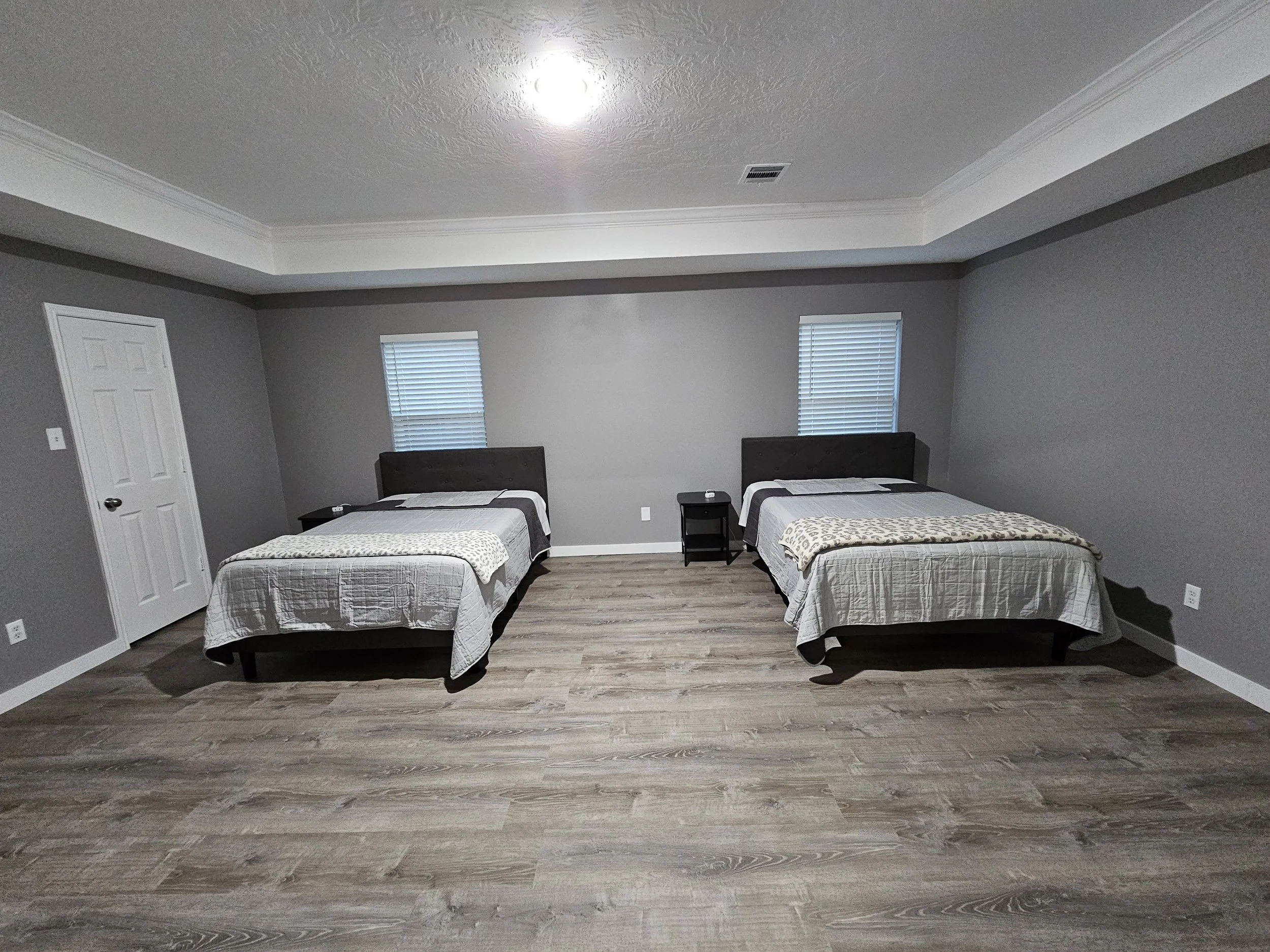 A bedroom with two single beds, each with a dark wooden headboard, white bedding, and a beige blanket. There is a small black nightstand between the beds and two windows with blinds on the back wall. The walls are painted light gray, and the floor ha