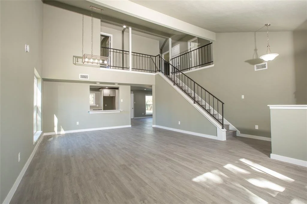 Empty living room with gray walls, large windows, and a staircase with black railings leading to an upper floor.