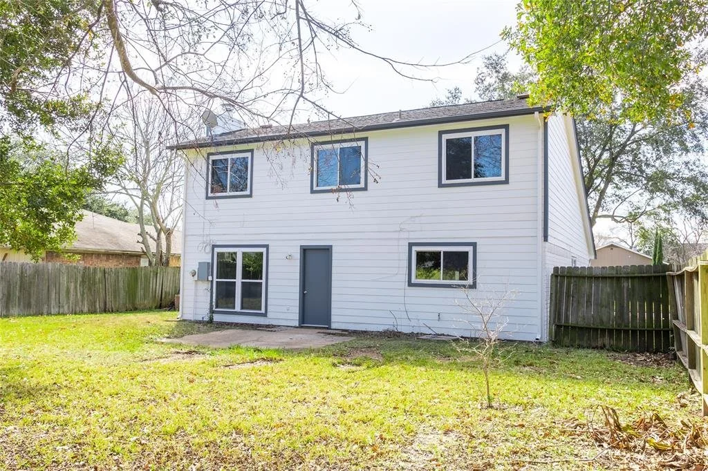 Back view of a two-story white house with black window frames, a gray door, and a fenced backyard with grass and a small tree.