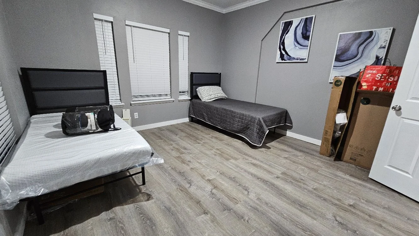 A room with two beds, gray walls, three windows with blinds, and three abstract art pieces on the wall. One bed has a plastic cover and a black bag on it, the other has a pillow and dark quilt. There are cardboard boxes and a plastic bag in the corne