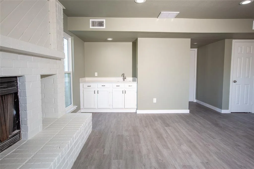 Empty living room with a white brick fireplace, a window, a white cabinet with a sink, and a door, featuring gray walls and hardwood flooring.