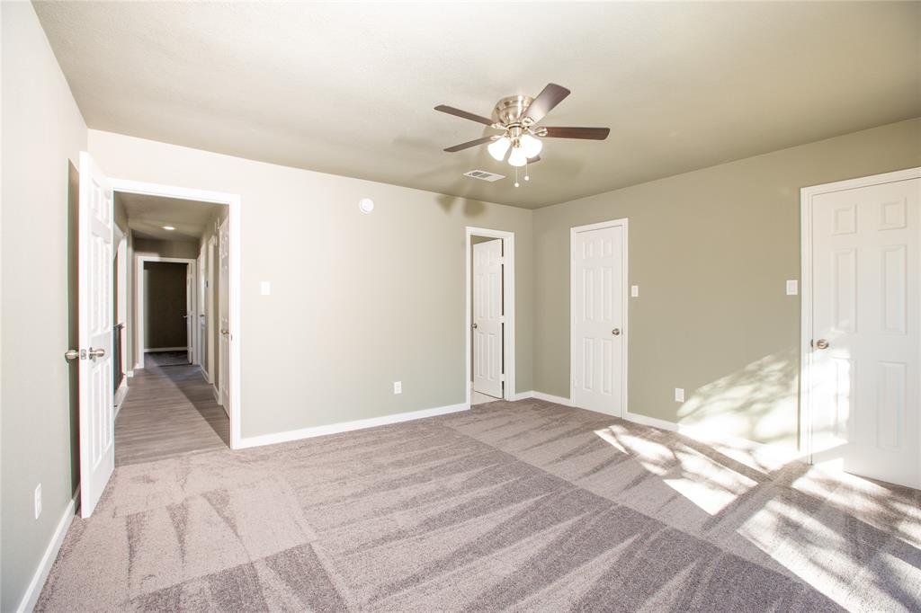 Empty room with beige carpet, light gray walls, four white doors, ceiling fan with lights, and sunlight coming through a window.