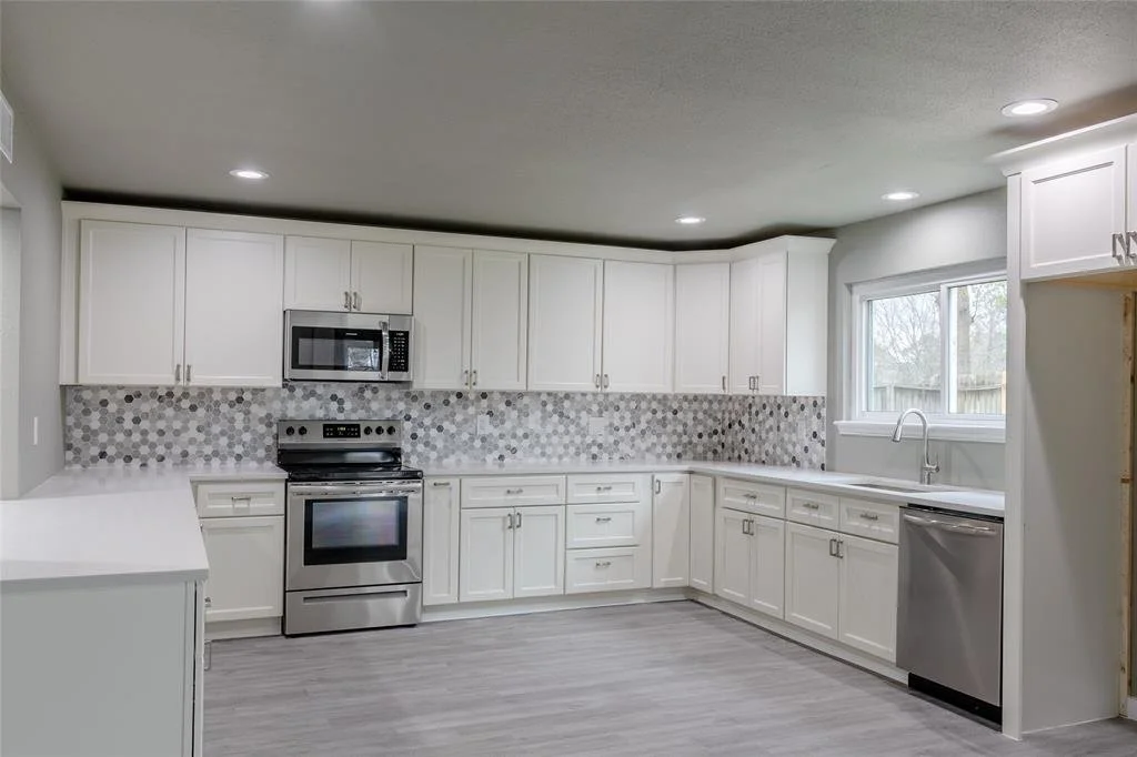Modern white kitchen with gray hardwood floors, white cabinets, gray and white backsplash, stainless steel appliances, a window over the sink, and recessed lighting.