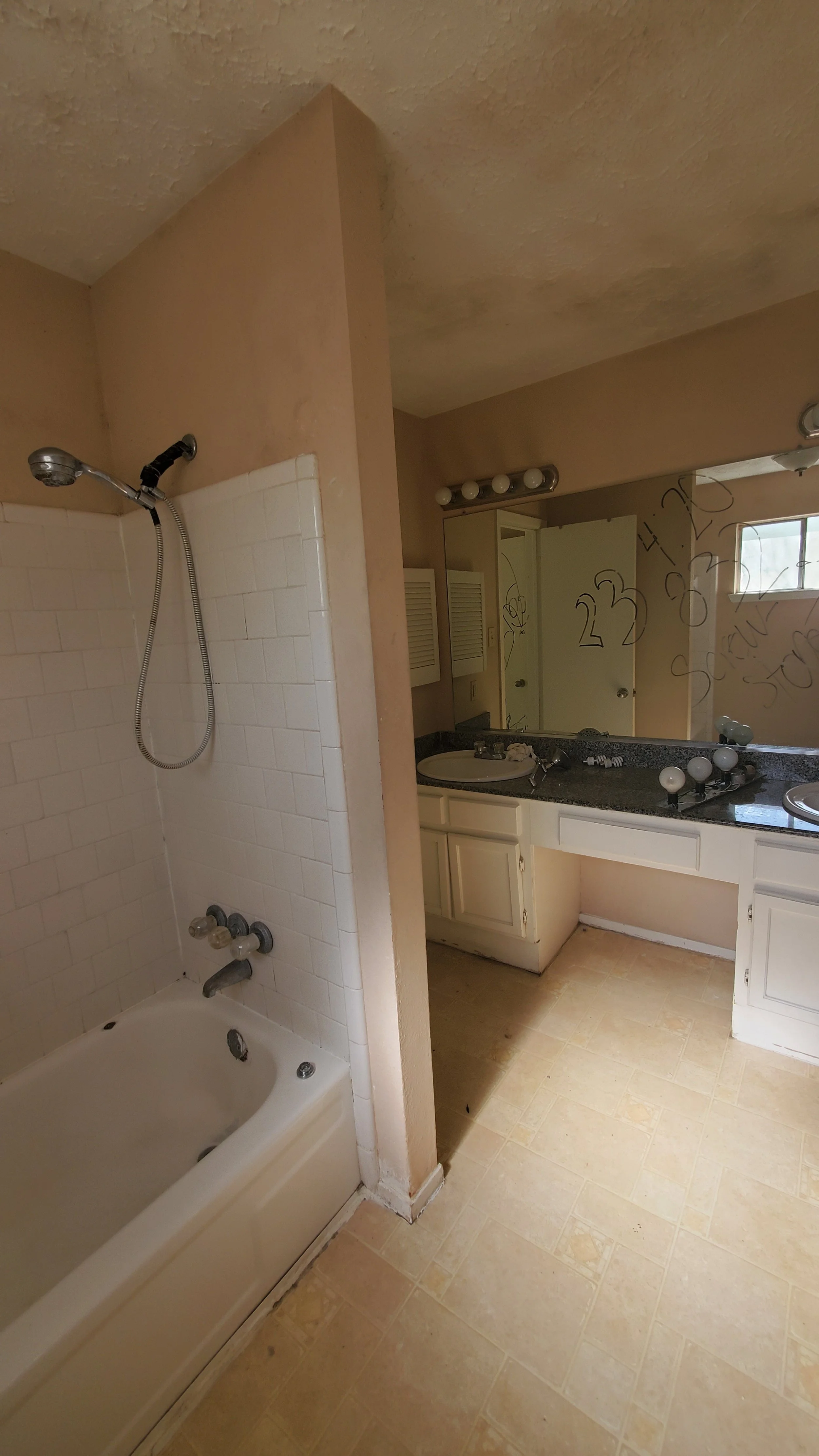 A bathroom with a bathtub, shower head, towel rack, and a vanity with a sink and mirror. The mirror has writing and markings on it, and the bathroom appears to be in a state of disrepair or renovation.