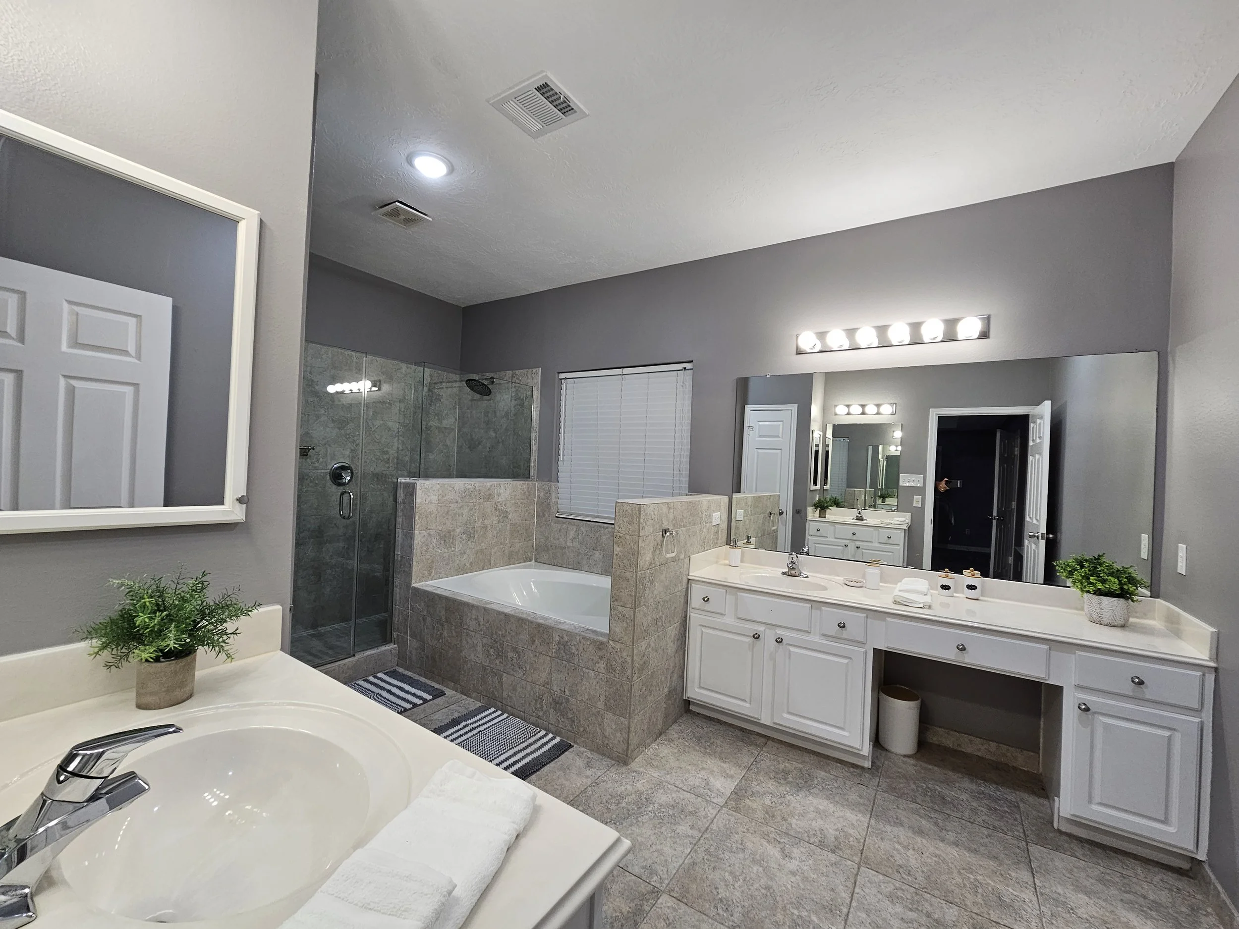 Modern bathroom with a large mirror, white vanity with a sink, potted plants, a bathtub with tile surround, glass-enclosed shower, gray painted walls, and tile flooring.