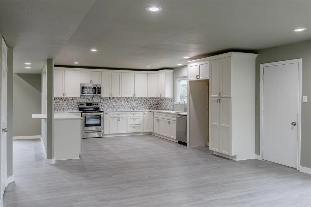 Empty modern kitchen with white cabinets, gray flooring, stainless steel appliances, and a patterned backsplash.