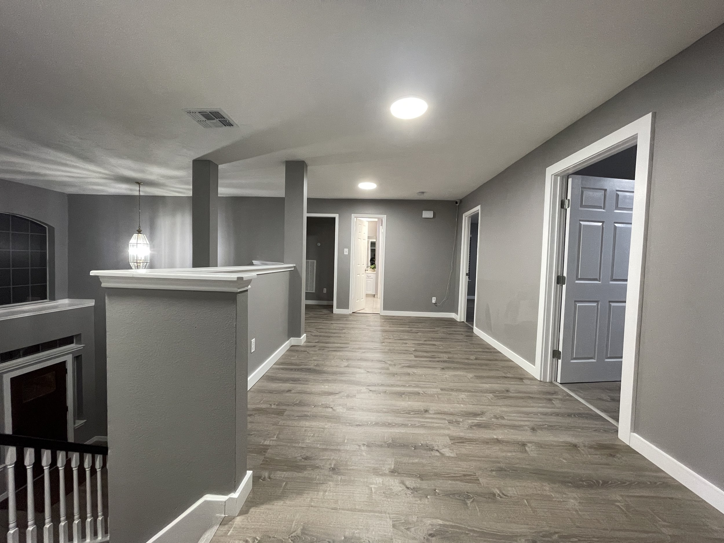 A spacious, modern upstairs hallway with gray walls, wood flooring, and multiple white-trimmed doorways leading to different rooms. There is a partial wall with a railing around a staircase, and ceiling lights illuminating the space.