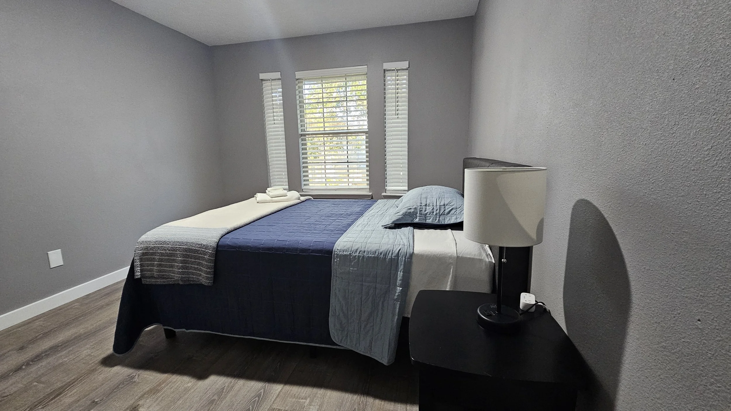 A bedroom with a bed covered with a blue and gray quilt, a pillow, and neatly folded towels at the head of the bed. Three windows with white blinds are behind the bed, allowing natural light into the room. To the right of the bed, there is a black ni