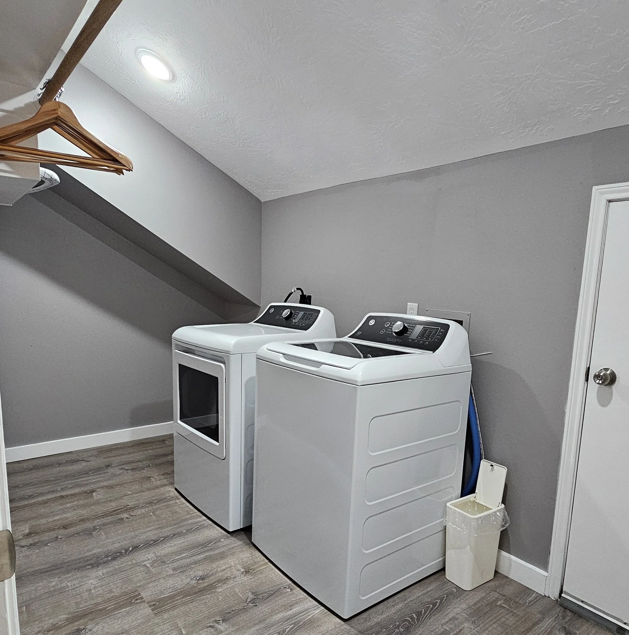 Laundry room with a white dryer and washing machine against a gray wall, a small trash bin with a lid next to the washing machine, a blue hose behind the machines, and a wooden wall-mounted hanger with clothes hangers on the left side.