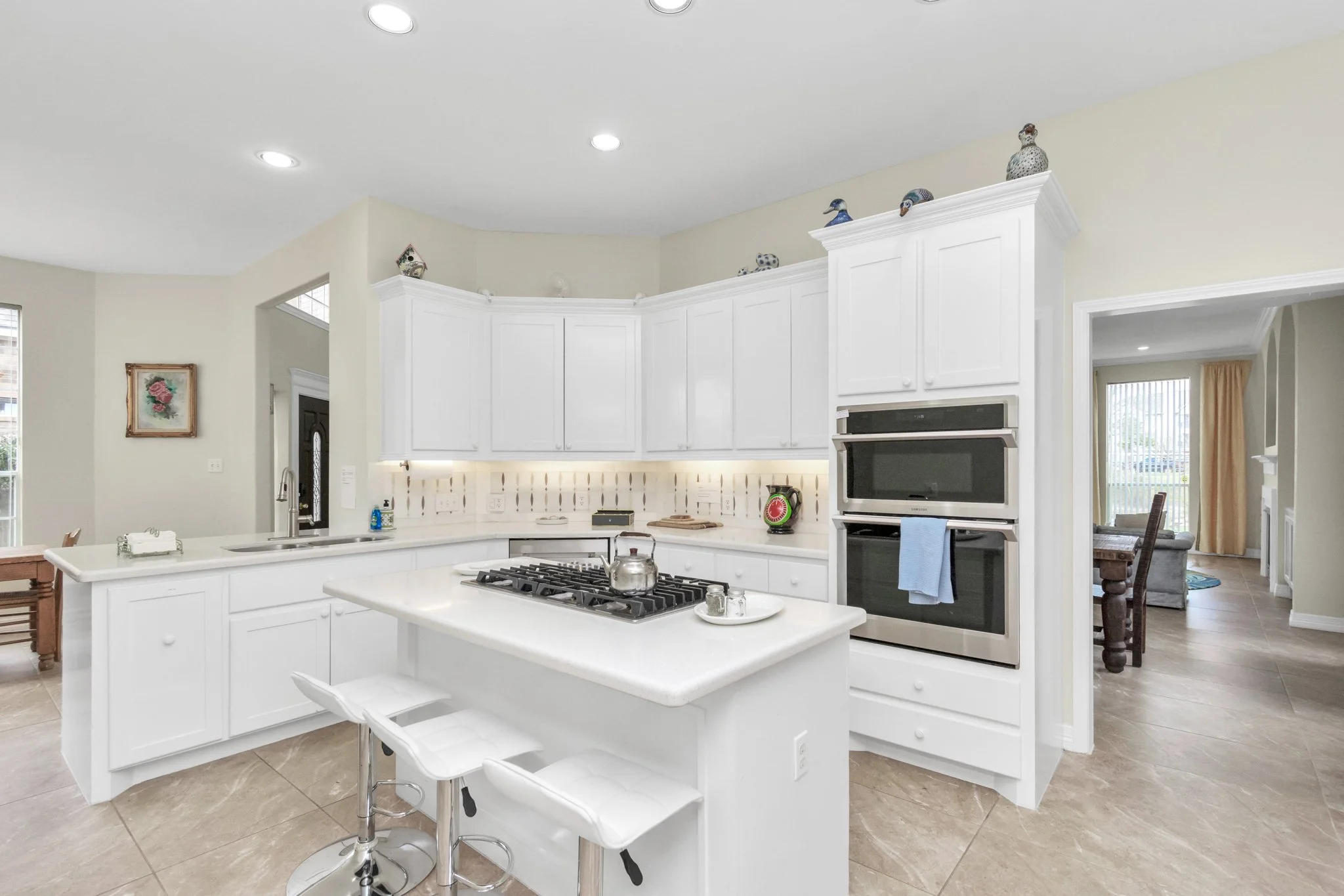 Bright kitchen with white cabinets, island with gas stove and tea kettle, double wall oven, and open doorway to a dining and living area.