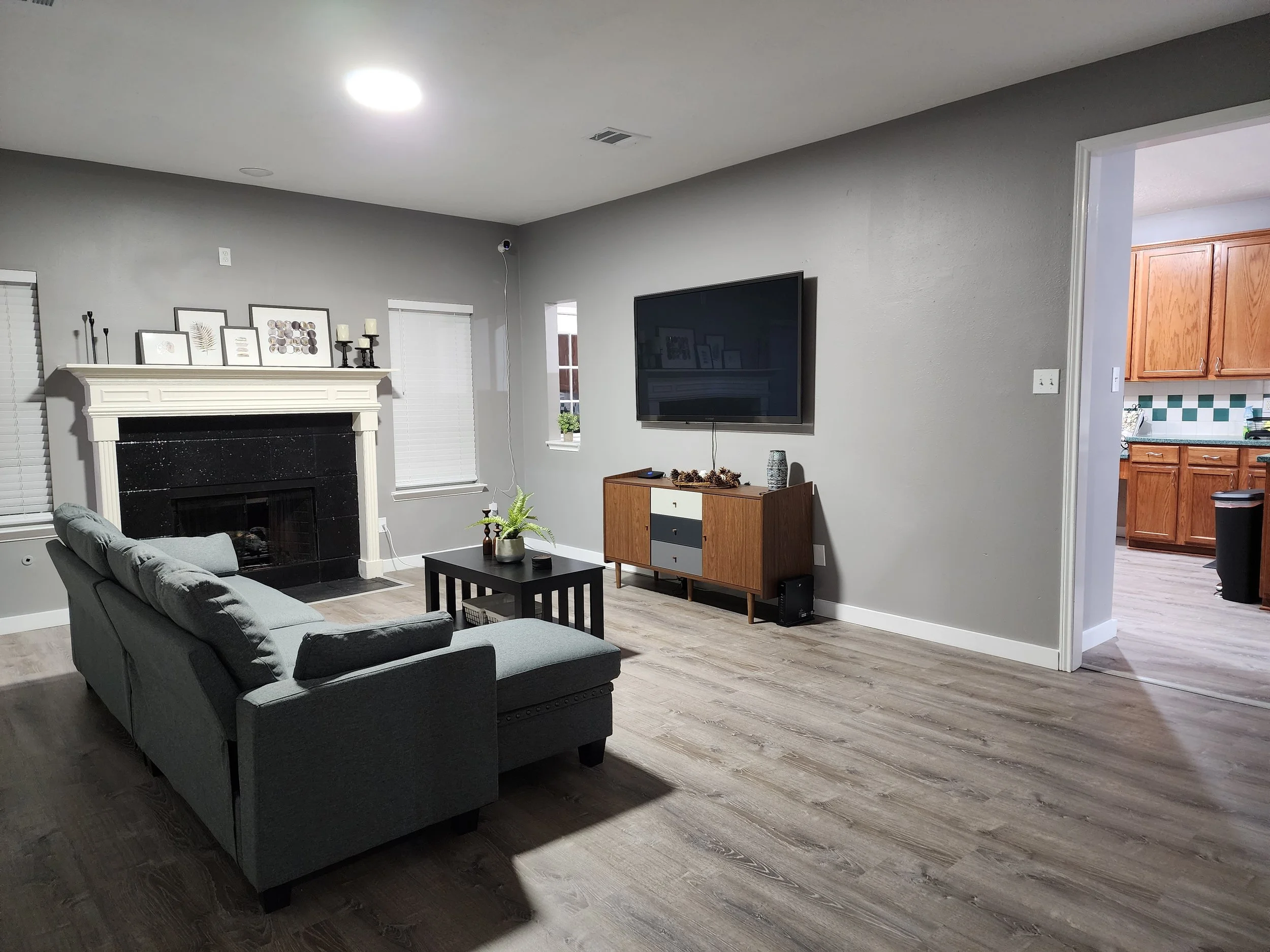 Living room with gray walls, a gray sofa, a black fireplace, and wall-mounted TV. There are windows with white blinds, a small side table with plant, and a TV stand with decor. An open doorway reveals a kitchen with wooden cabinets.
