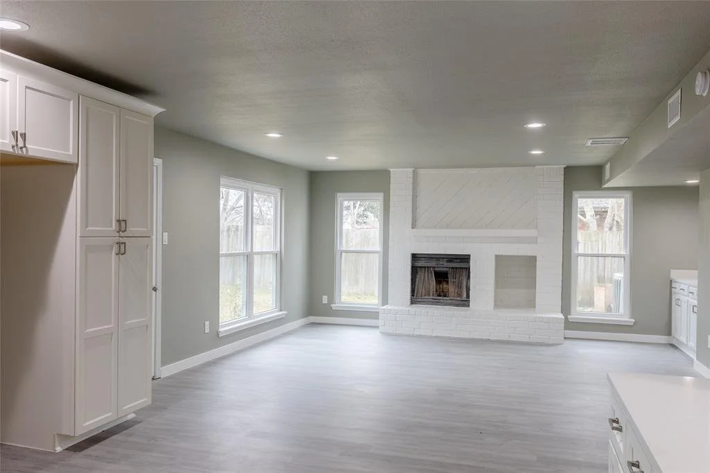 Bright living room with large windows, white painted brick fireplace, light-colored hardwood floors, and white kitchen cabinets.
