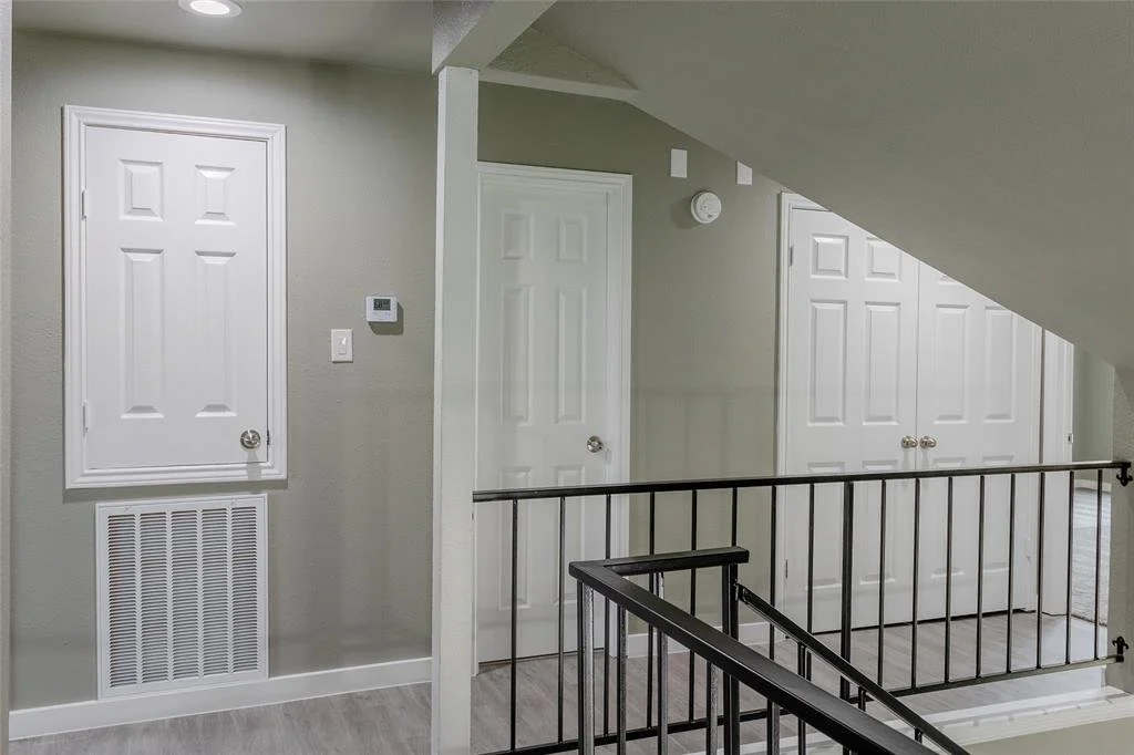 Interior view of a house hallway with four closed white doors, a smoke detector, thermostat, and a vent on the wall, with a staircase and black metal railing.