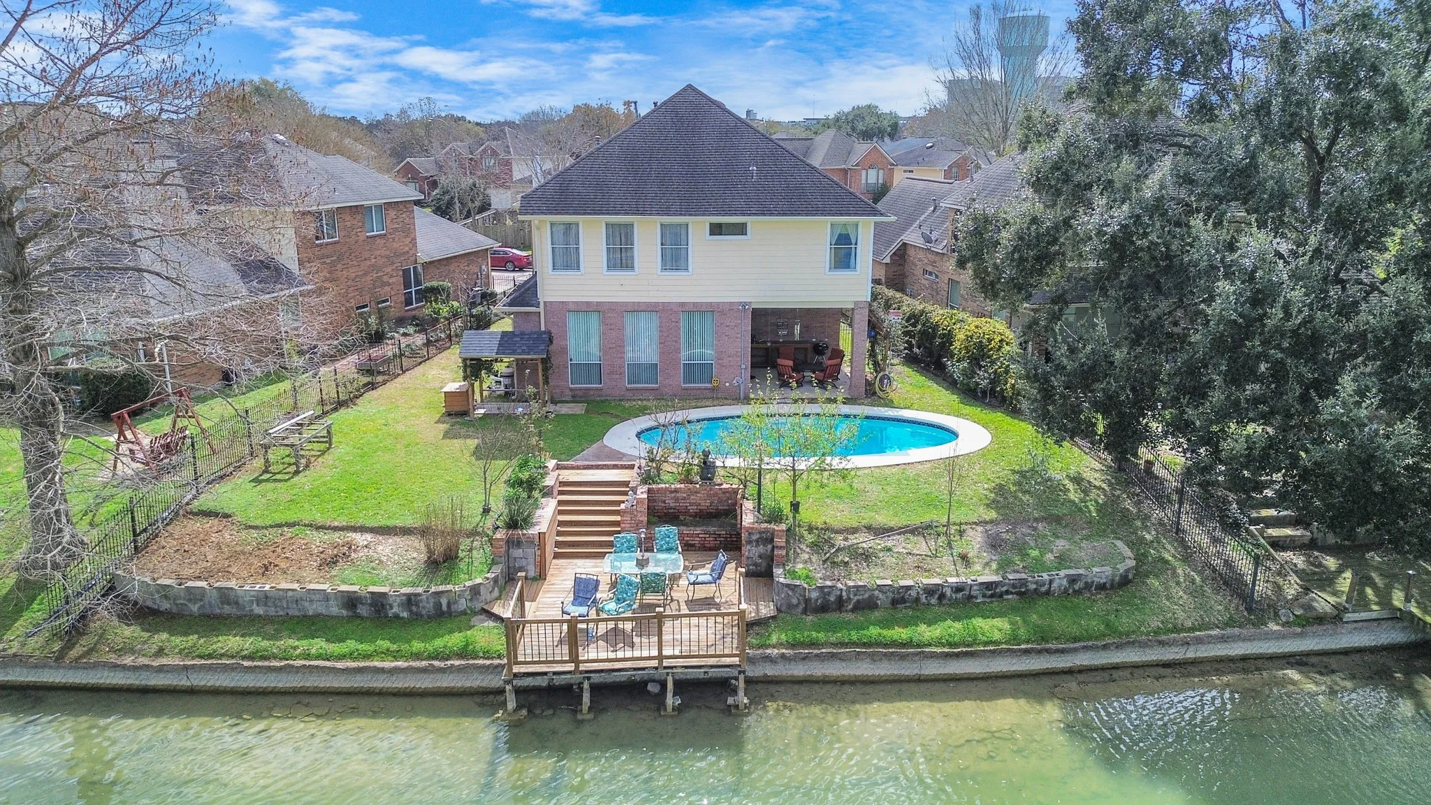 Aerial view of a two-story house with a backyard that includes a swimming pool, outdoor patio with chairs, and a dock along a waterway, surrounded by trees and neighboring houses.