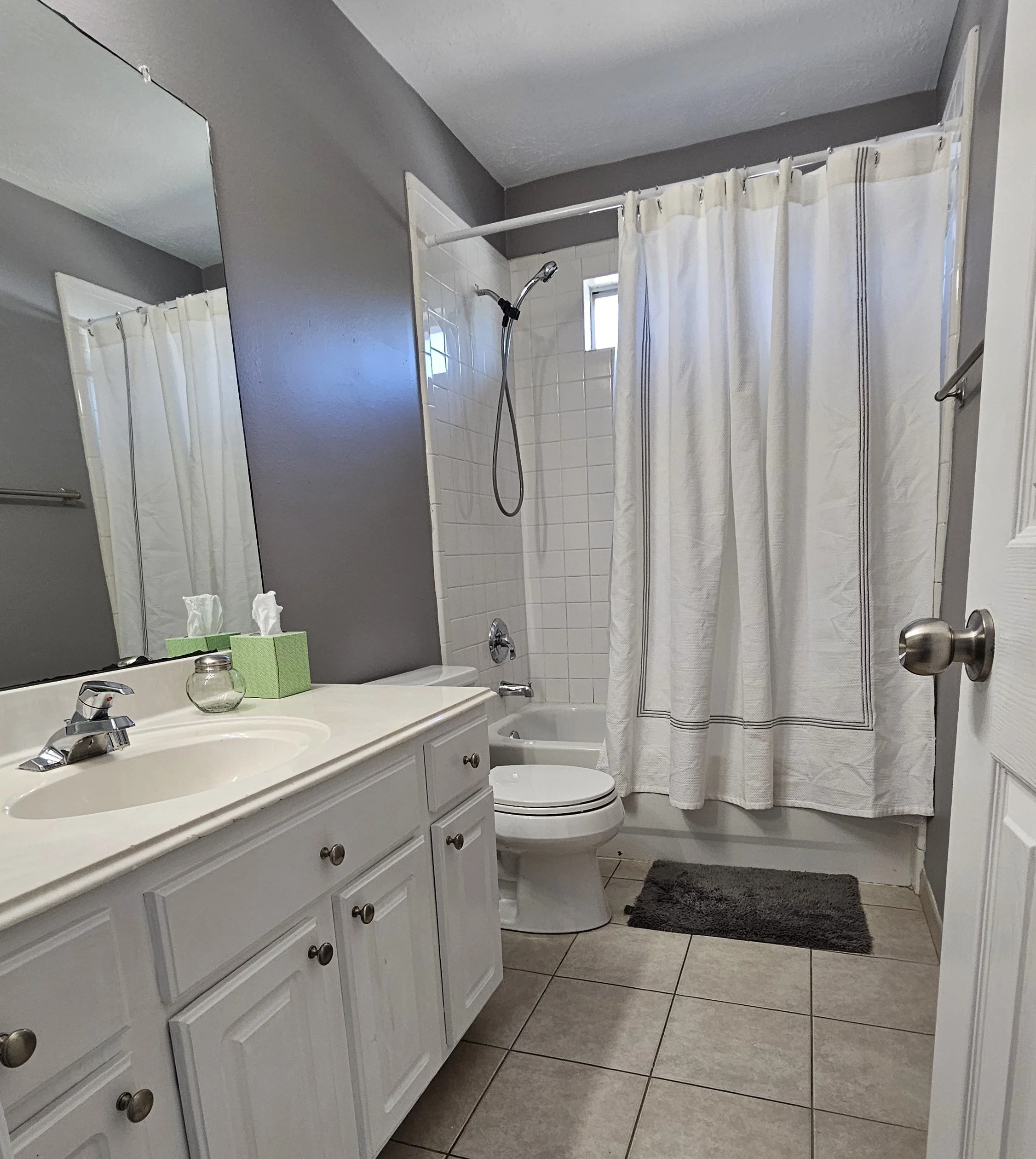 A small bathroom with a white vanity sink, a mirror, a toilet, and a shower with a curtain. There are tissue boxes and a small jar on the vanity, gray walls, and a gray rug on the tiled floor.