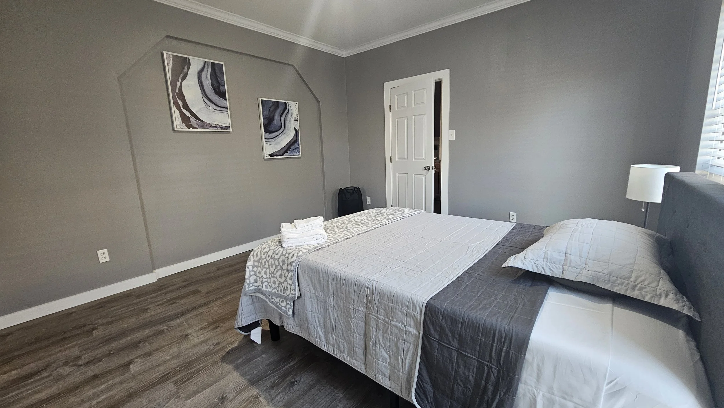 A bedroom with a gray wall, a bed with gray and white bedding, a pillow, a nightstand with a lamp, and two abstract wall art pieces. The floor has wood-like flooring, and a window with blinds is on the right side.
