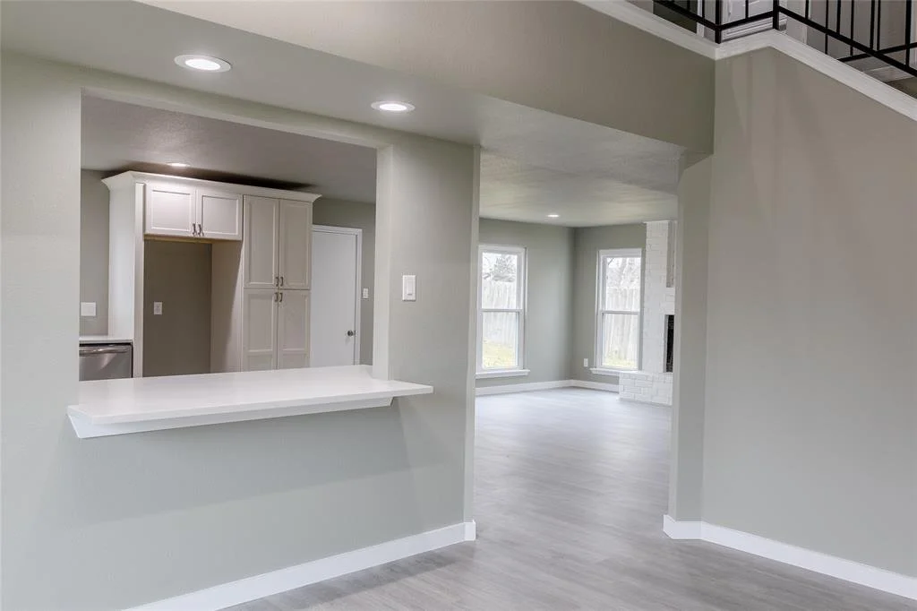 Empty living room and kitchen area in a modern home with light gray walls, white cabinetry, large windows, and light wood flooring.