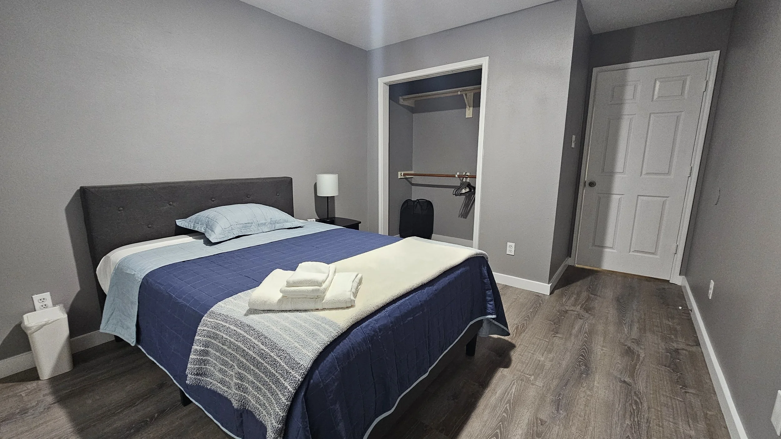 A bedroom with a gray upholstered bed, blue bedding, and a gray pillow. A nightstand with a lamp is beside the bed. An open closet with a black bag and hanging space is in the background. There are folded towels on the bed.