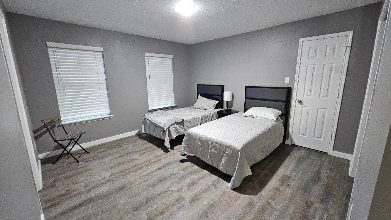 A bedroom with two twin beds, each with white bedding and located next to each other. There are two windows with white blinds, a small side table with a lamp between the beds, and a folding chair near the wall. The room has gray walls and wood floori