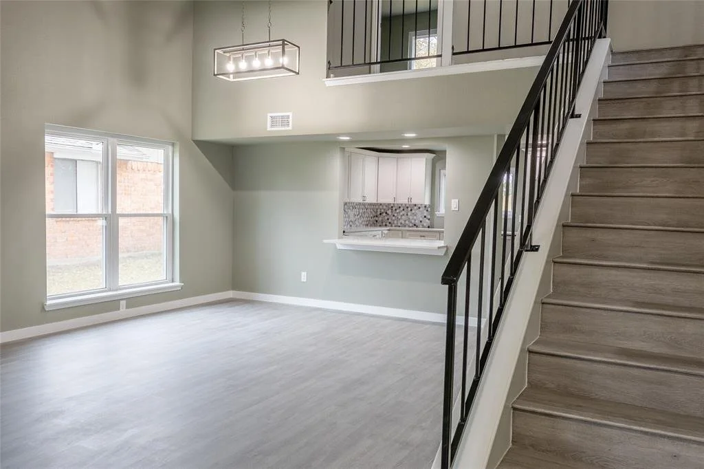 Empty living room with large window, modern chandelier, light-colored flooring, and a staircase leading to an upper level with a hallway view of a kitchen in the background.