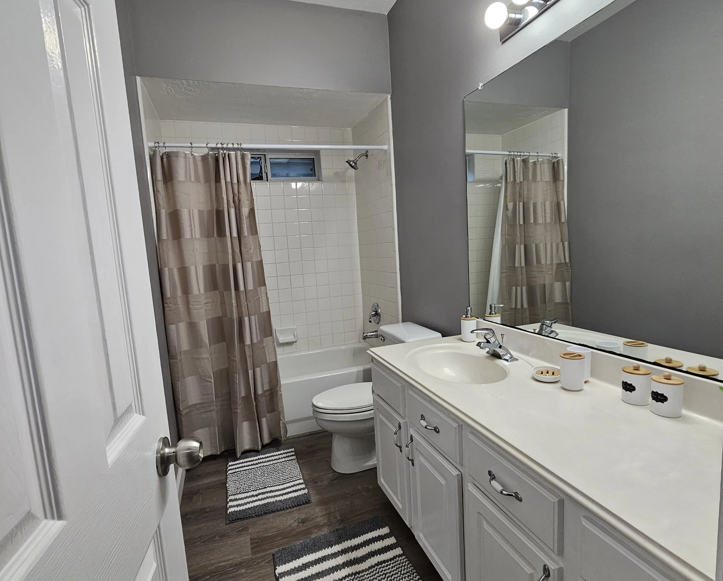 A bathroom with a white vanity, large mirror, and gray walls. The vanity has a sink with a chrome faucet, and various soap dispensers and containers. To the left, there is a toilet and a bathtub with a beige striped shower curtain. The shower area ha