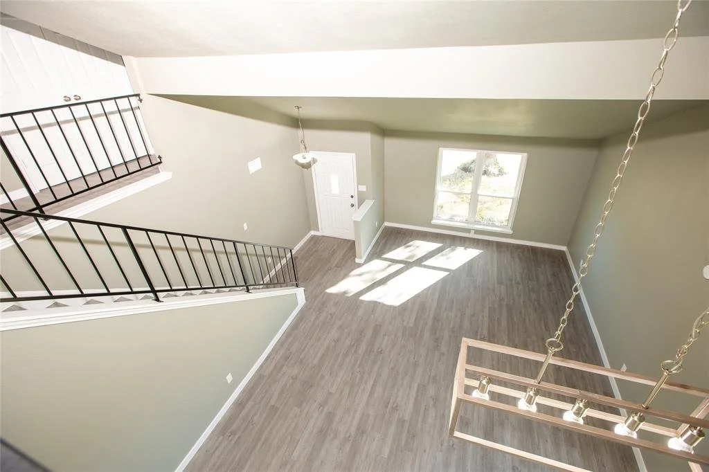 Empty living room with light gray walls, wood flooring, a large window, and a black metal staircase railing.