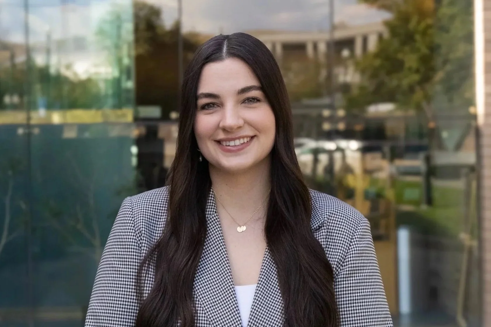 A young woman with long dark hair, smiling, standing outdoors in front of a glass building with trees and a bridge reflected in the glass.