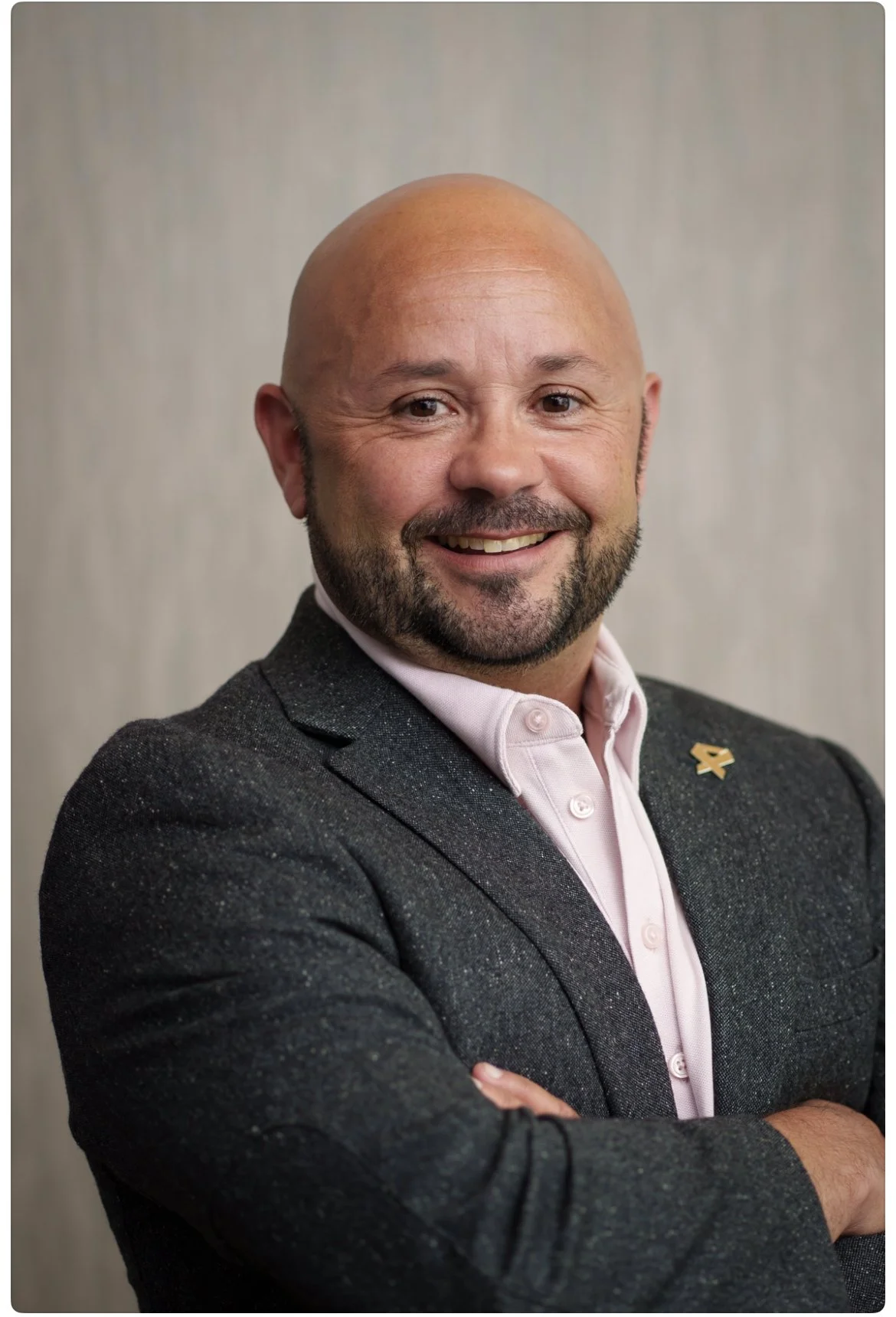 A smiling bald man with a beard and mustache, wearing a dark suit jacket and a light pink dress shirt, standing against a plain background, with a small ribbon pin on his jacket lapel.