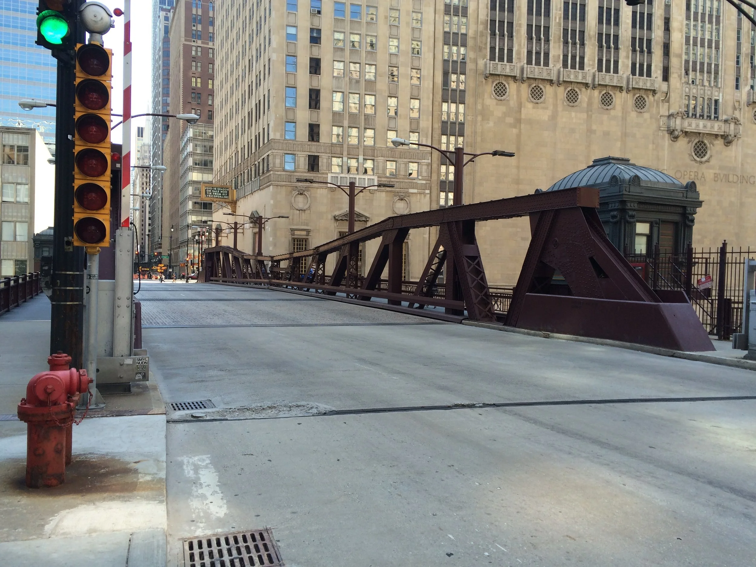 Empty city street with a traffic light showing green, a fire hydrant, tall buildings, and a rusty bridge in the background.