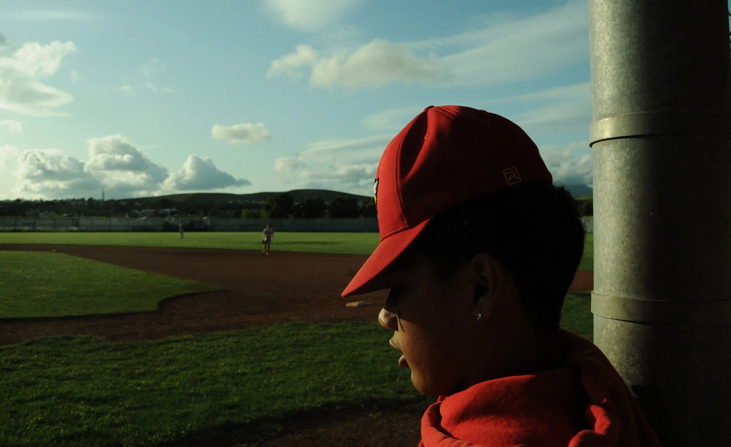A woman wearing a red baseball cap and earrings standing near a baseball field, with the field and players visible in the background under a partly cloudy sky.