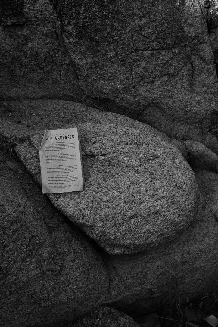 A crumpled piece of paper with printed text sitting on a large smooth rock among other rocks in a natural outdoor setting.